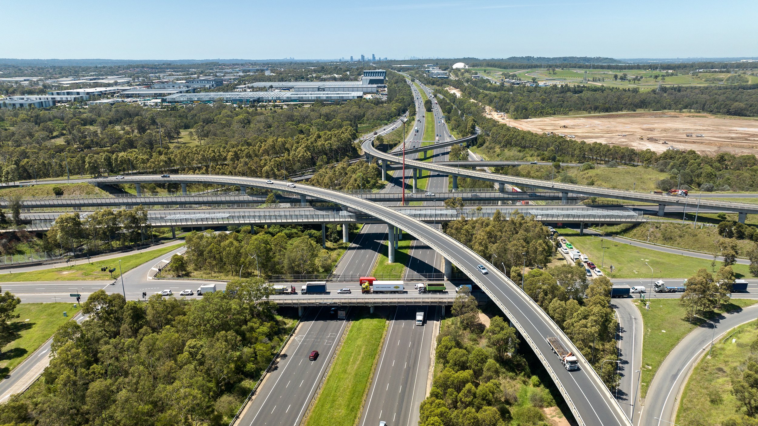 Aerial view of multiple highways and overpasses with cars, trucks, and greenery, leading toward a city skyline in the distance.