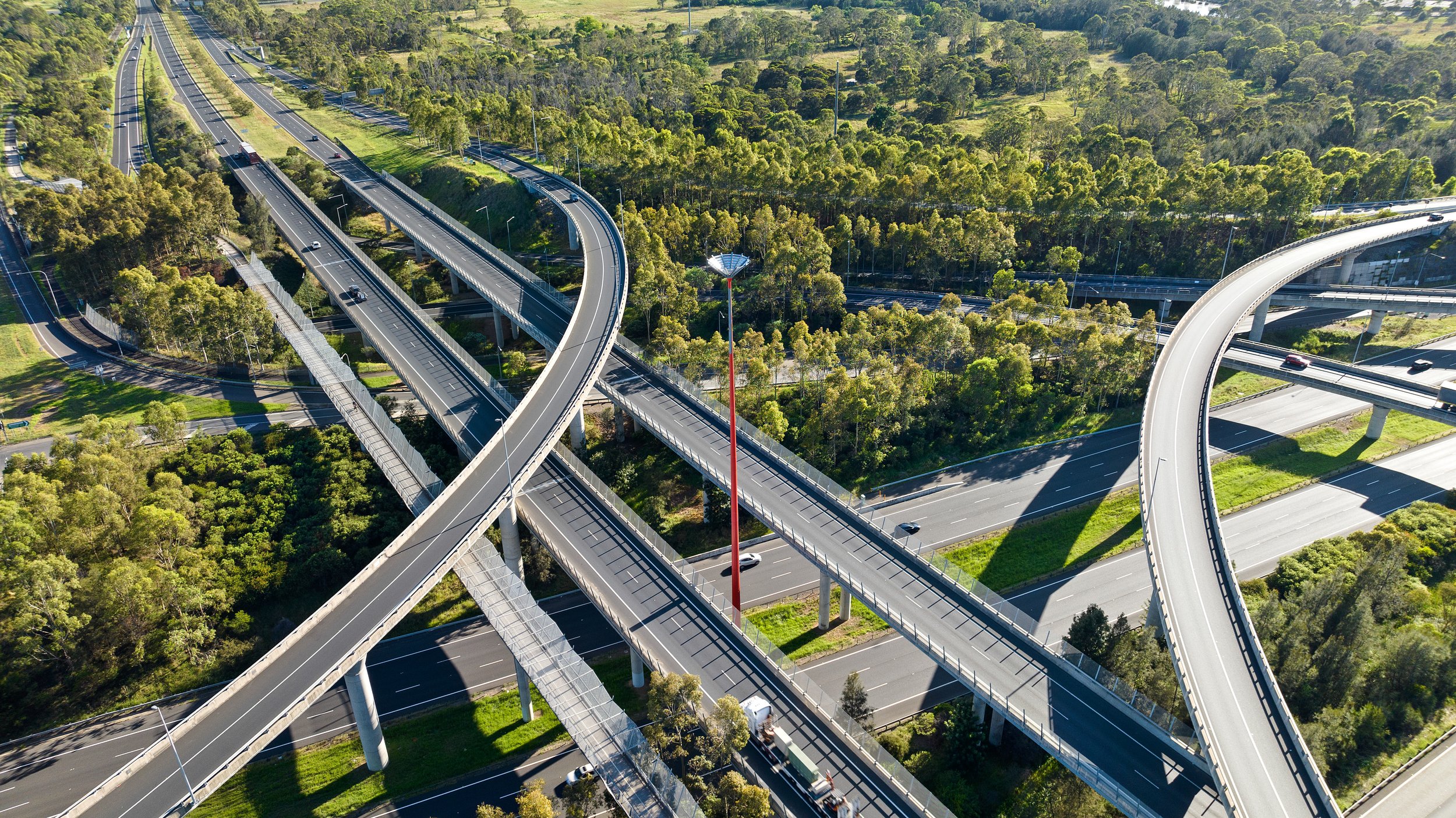 An aerial view of a complex highway interchange with multiple overpasses and bridges, surrounded by green trees and foliage. Designed by Colin Polwarth Studio Photo by Ruth Gold