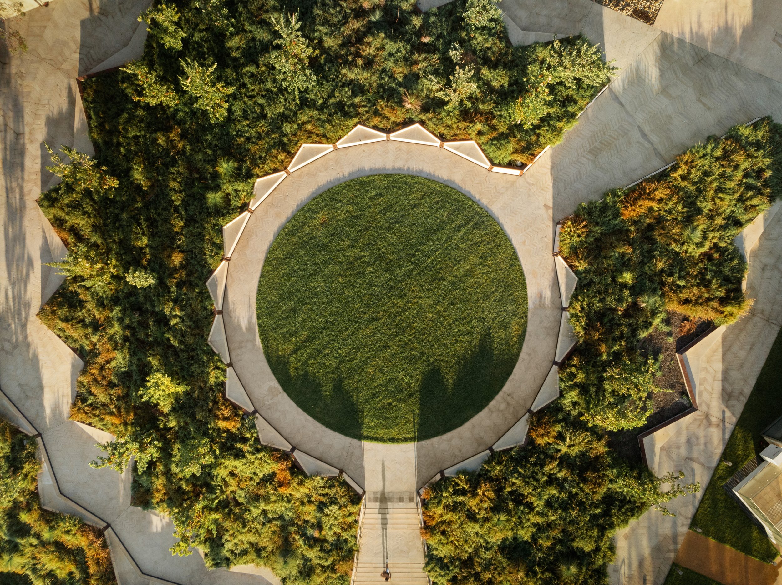 Aerial view of a landscaped garden with a circular grassy area surrounded by curved walkways and lush trees. Designed by McGregor Coxall Photo by Ruth Gold
