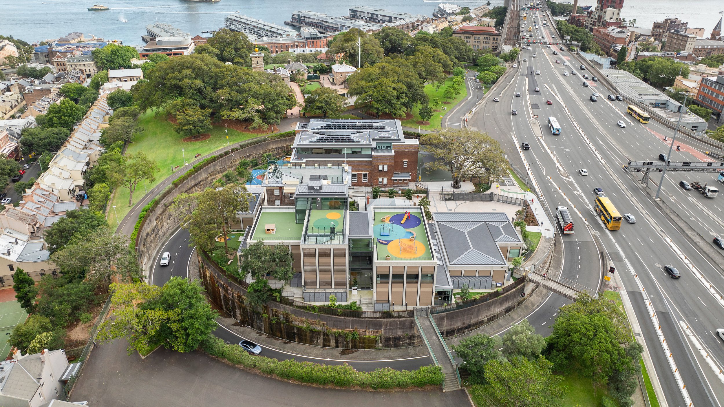 Aerial view of a modern building with colorful rooftop play area, surrounded by urban landscape including a highway, residential houses, and green park. Designed by fjcStudio Photo by Ruth Gold