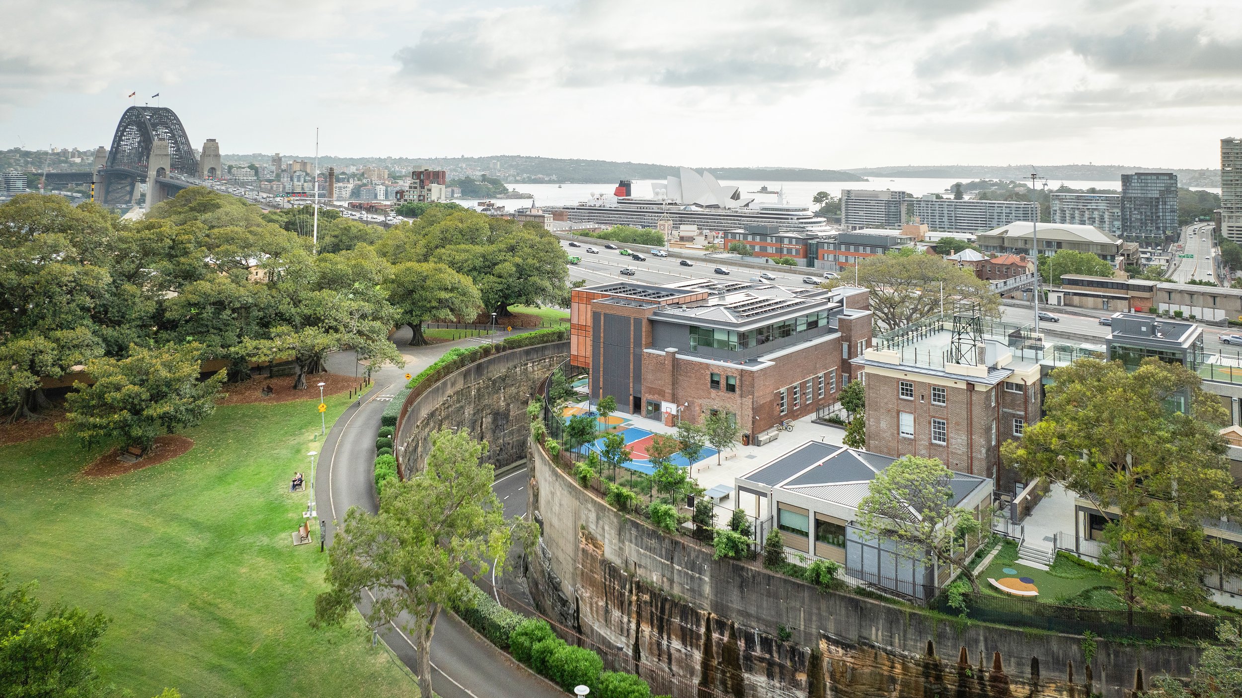 View of a cityscape with a park, a curved road, and buildings, including the Sydney Opera House, in the background. Designed by fjcStudio Photo by Ruth Gold