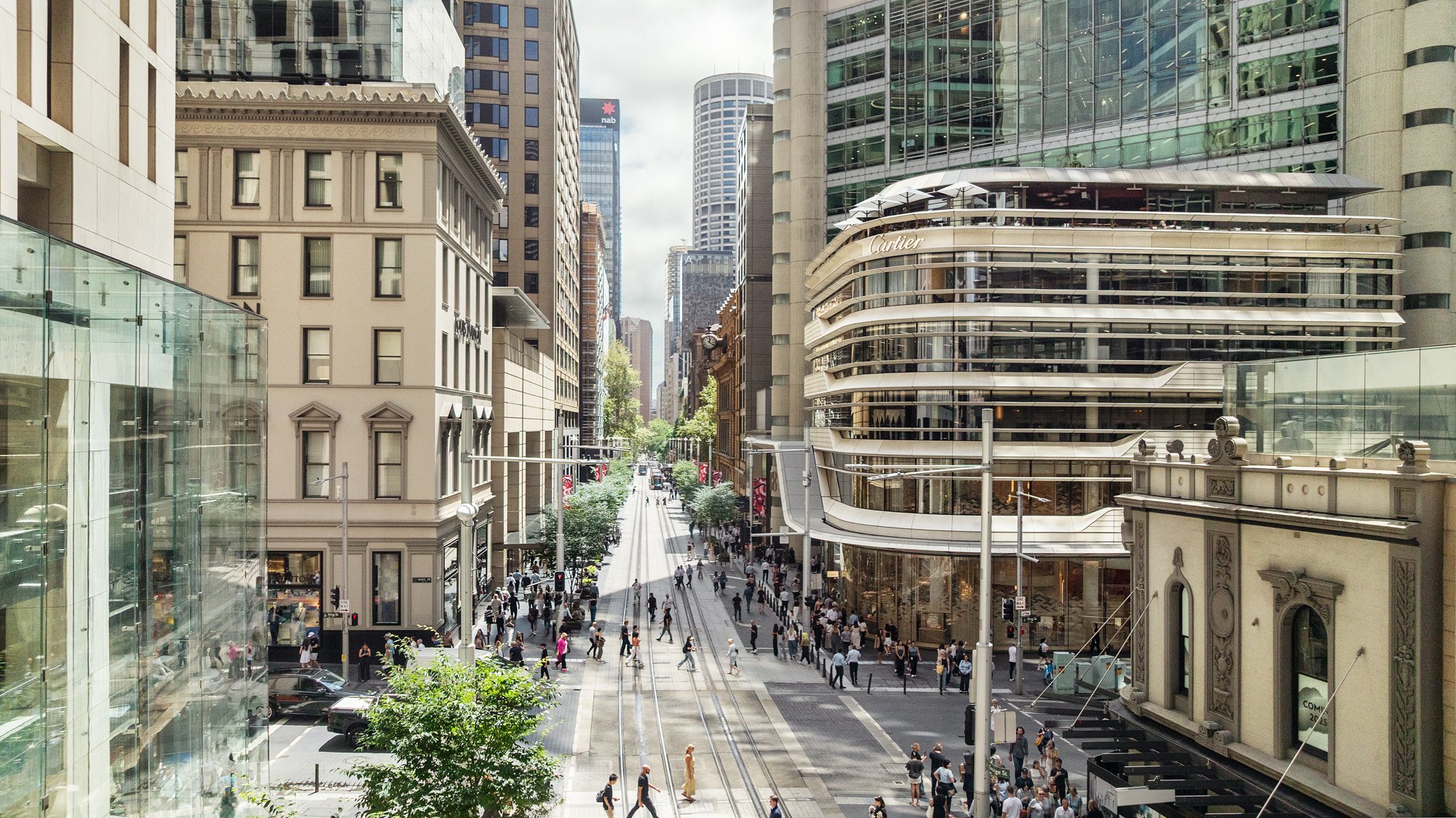 City street scene with high-rise buildings, pedestrians crossing the street, and tram tracks. Designed by fjcStudio Photo by Ruth Gold
