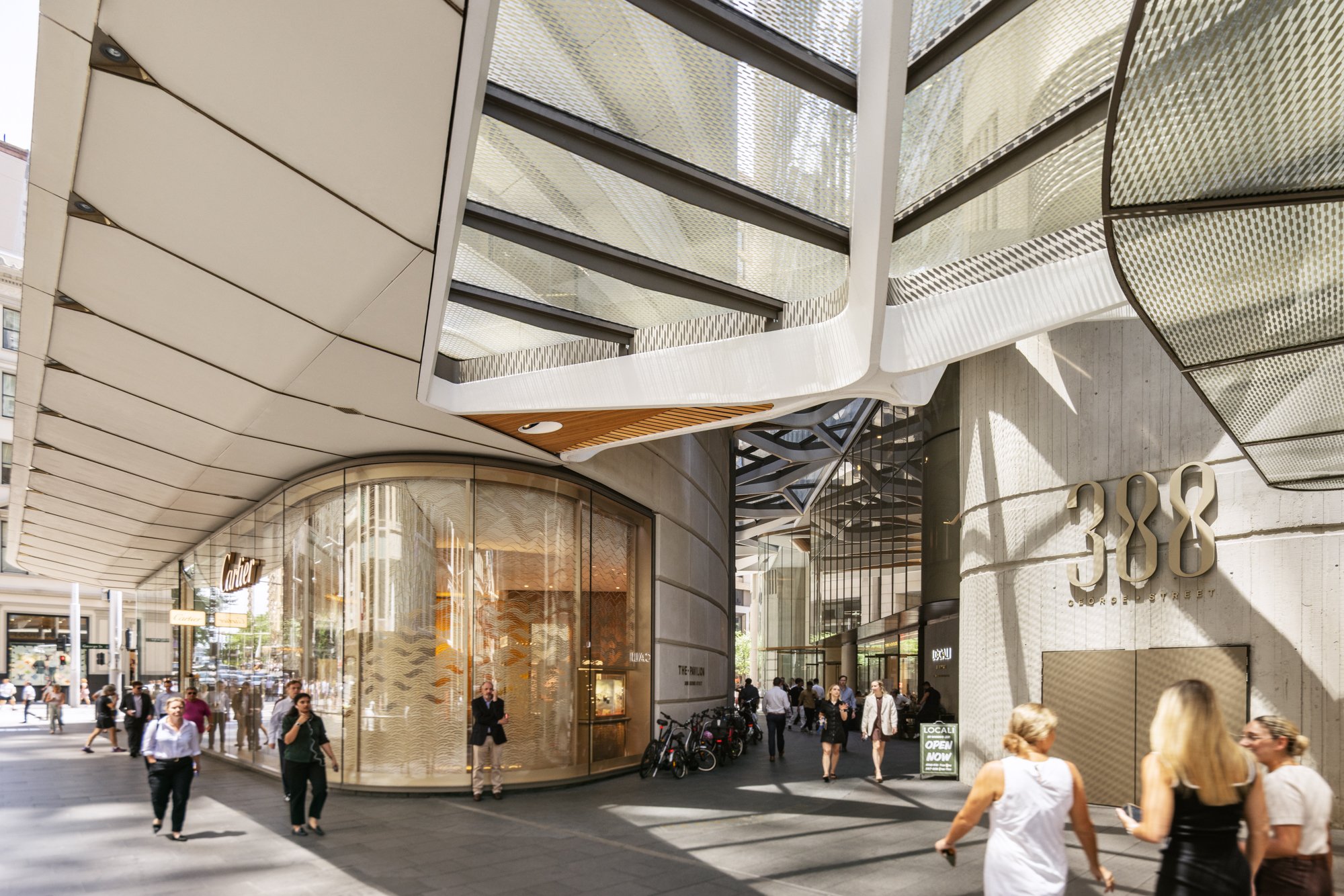 Modern shopping mall entrance with large glass windows, a curved facade, and the address 388 on the wall. Several pedestrians walking and standing outside, some talking and taking photos. Bright sunlight creating shadows on the ground. Designed by fj