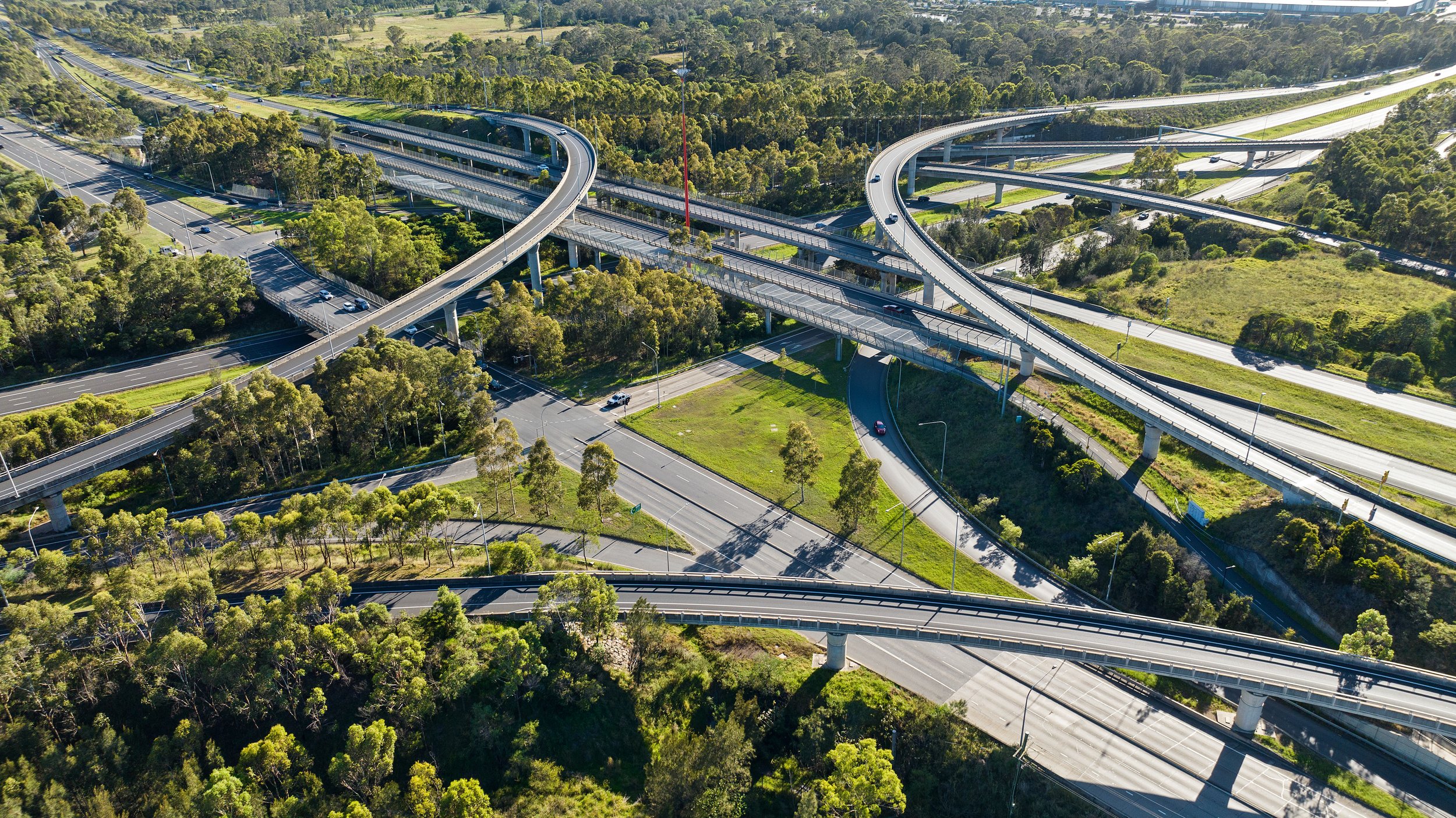 An aerial view of a complex highway interchange with multiple elevated roads and overpasses surrounded by green trees and open land. Designed by Colin Polwarth Studio Photo by Ruth Gold
