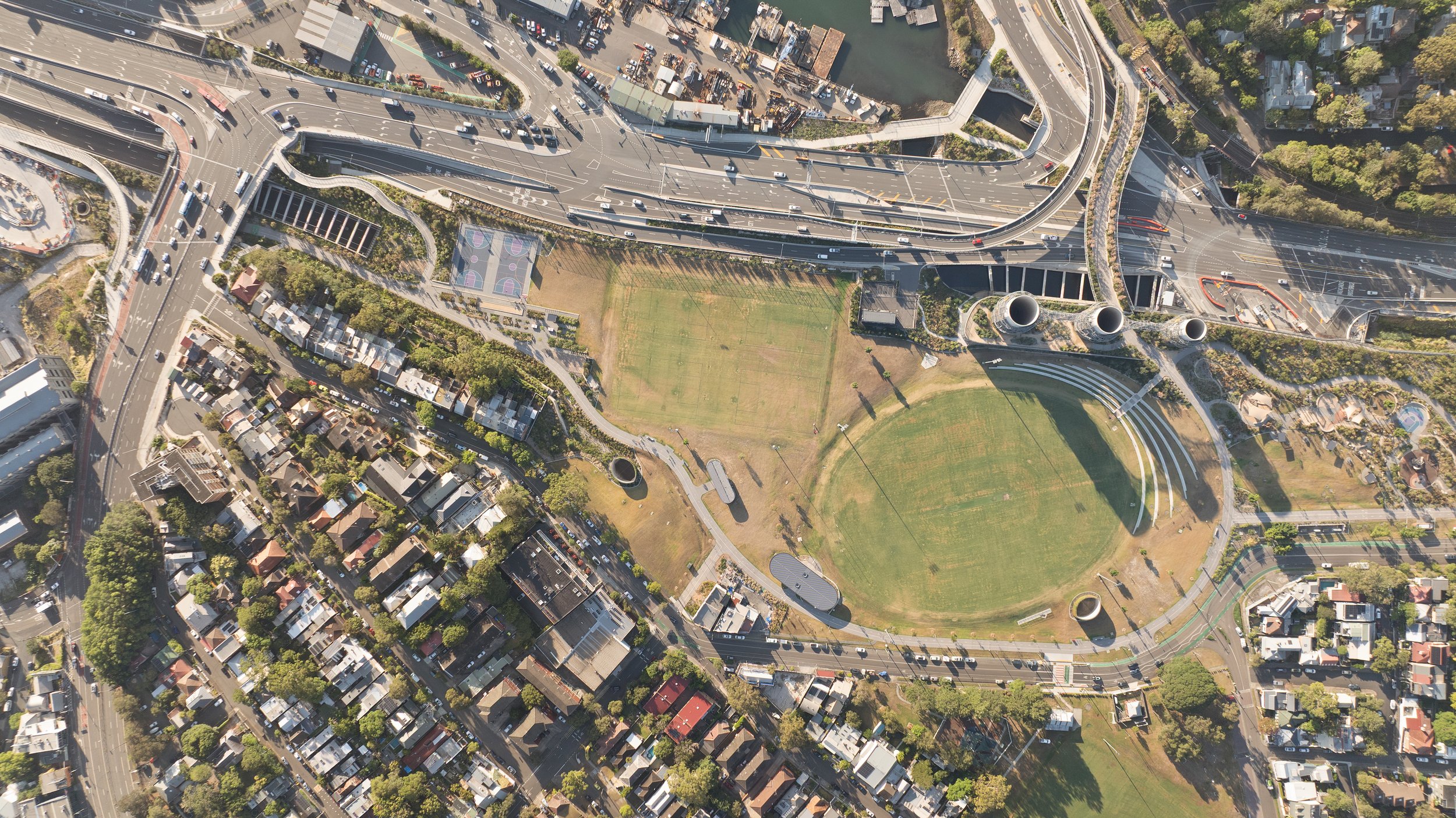An aerial view of a city park with open grass fields, surrounding houses, roadways, and overpasses, with some traffic visible. Designed by McGregor Coxall Photo by Ruth Gold