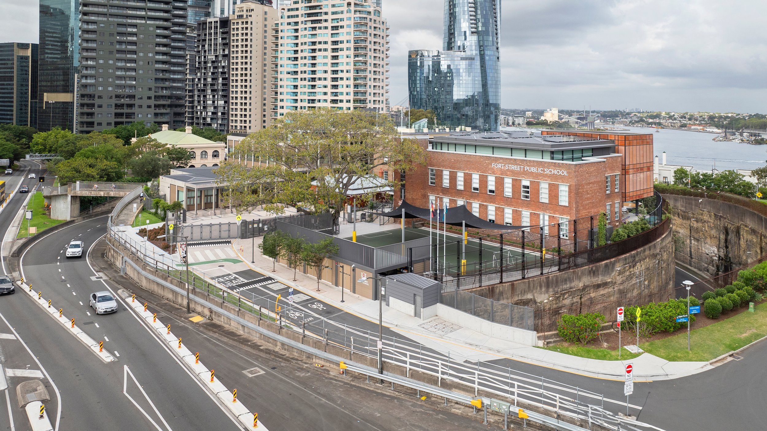 A cityscape featuring a school building labeled 'Fort Street Public School', surrounded by high-rise buildings, a river in the background, and busy roads with traffic. Designed by fjcStudio Photo by Ruth Gold