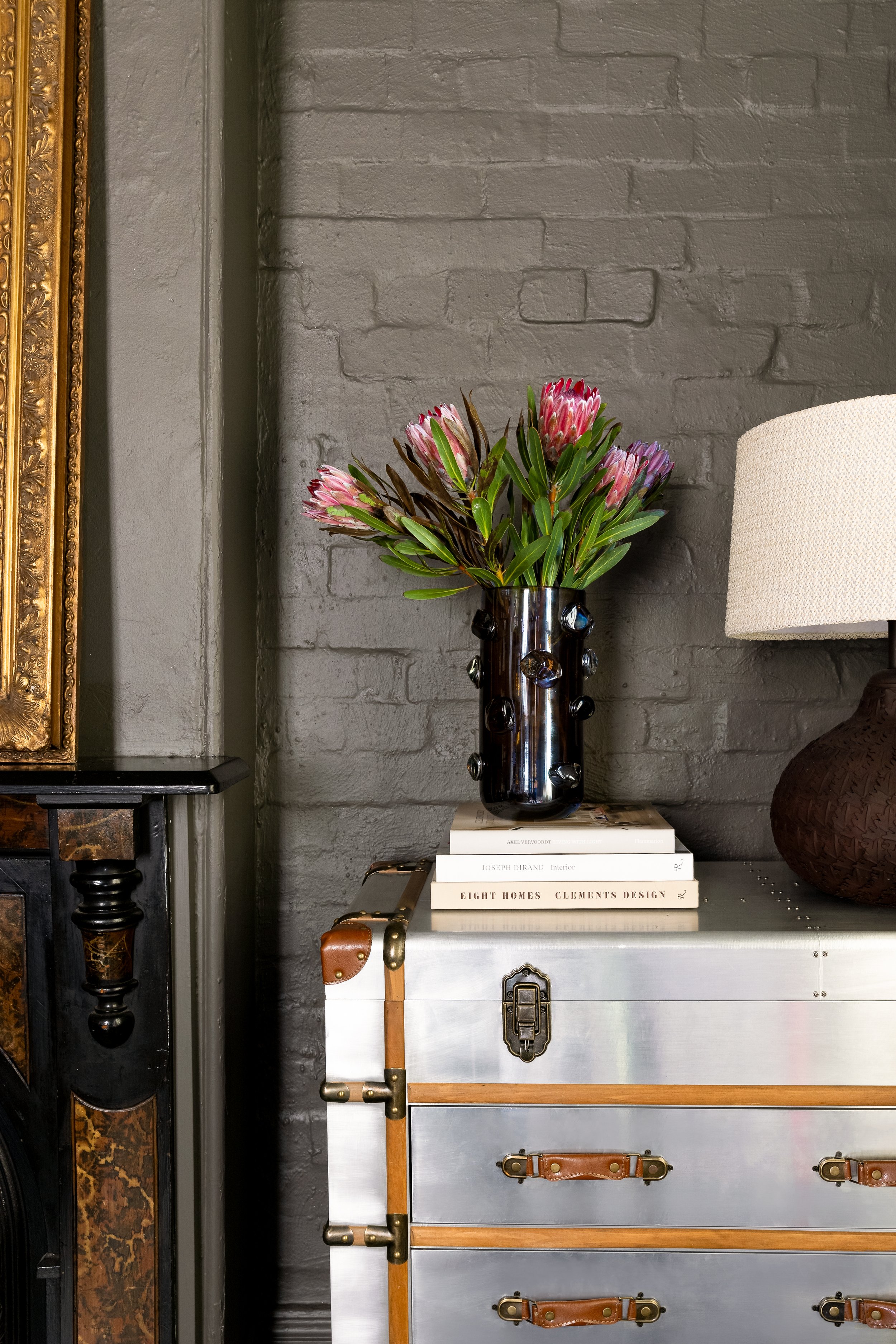 A black vase with decorative stones contains pink protea flowers with green leaves, placed on a stack of interior design books on a metallic dresser with leather accents, against a gray brick wall, next to a table lamp. SMAC Studio Photo by Ruth Gold