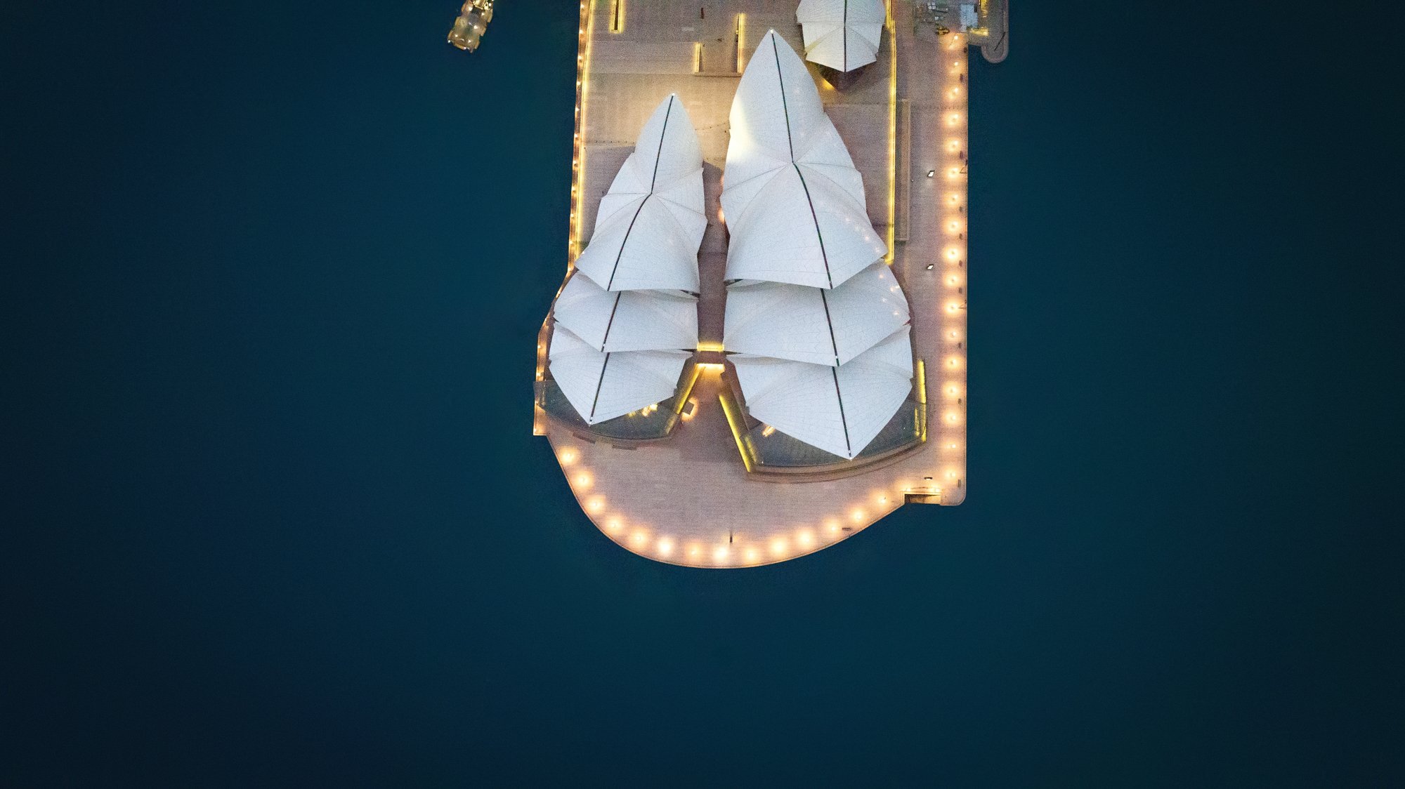 Aerial view of a rooftop with white, tent-like structures surrounded by string lights on a waterfront. Opera House Photo by Ruth Gold