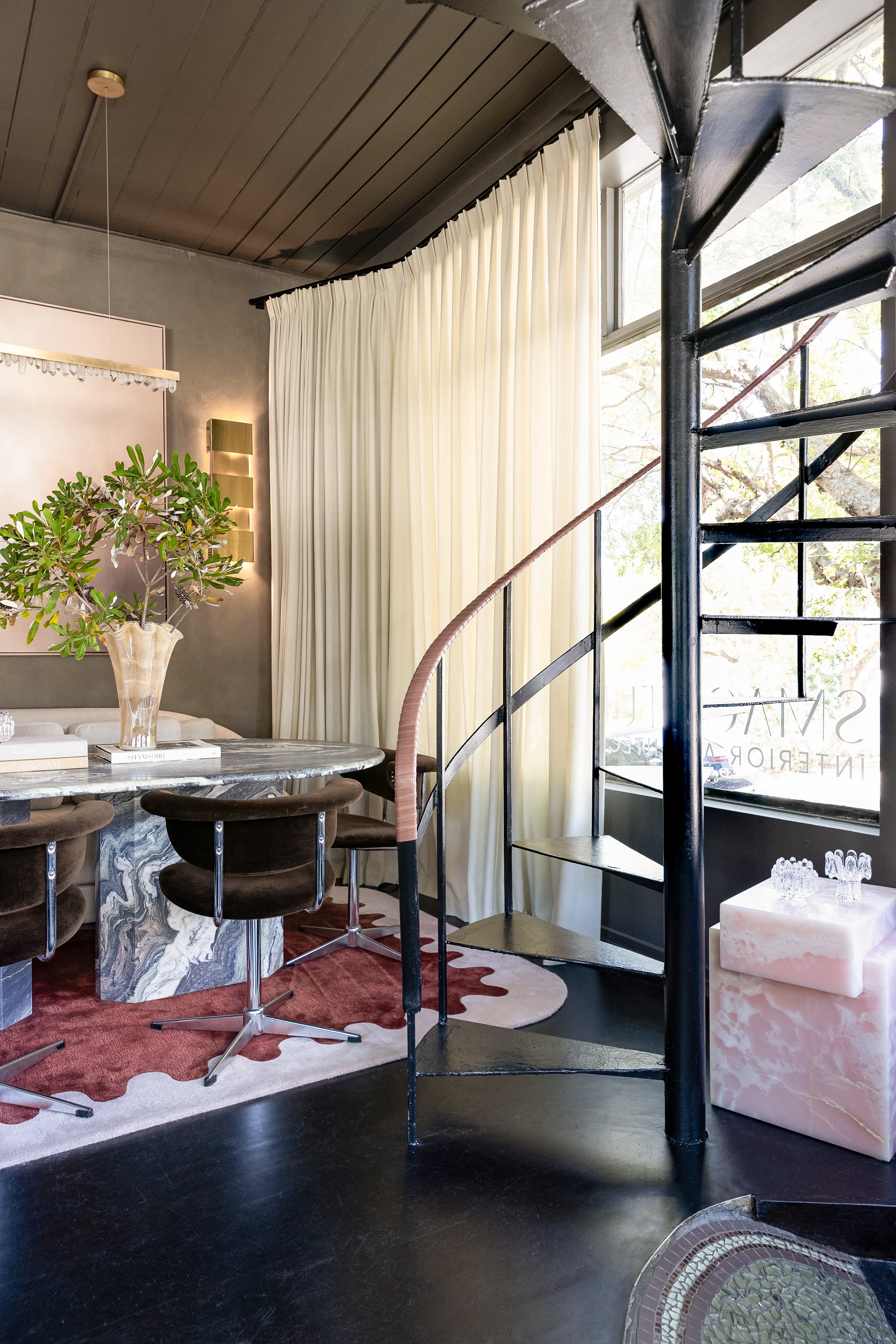 Interior of a modern living space with a round marble table, dark brown chairs, a large plant, cream curtains, a spiral staircase, and large windows letting in natural light. SMAC Studio Photo by Ruth Gold