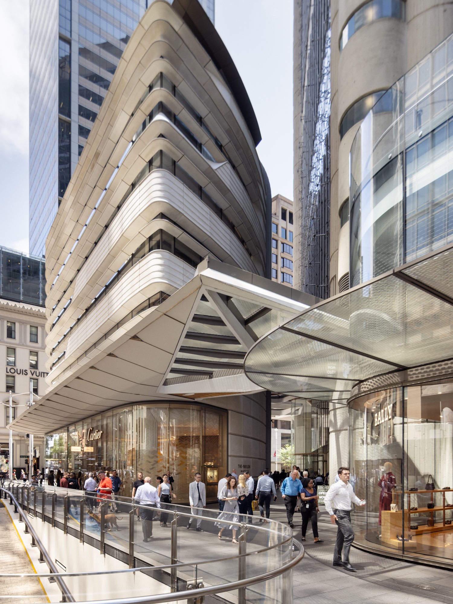 City street scene with modern high-rise buildings, a shopping mall entrance with people walking, and storefronts for luxury brands like Cartier and Bally. Designed by fjcStudio Photo by Ruth Gold
