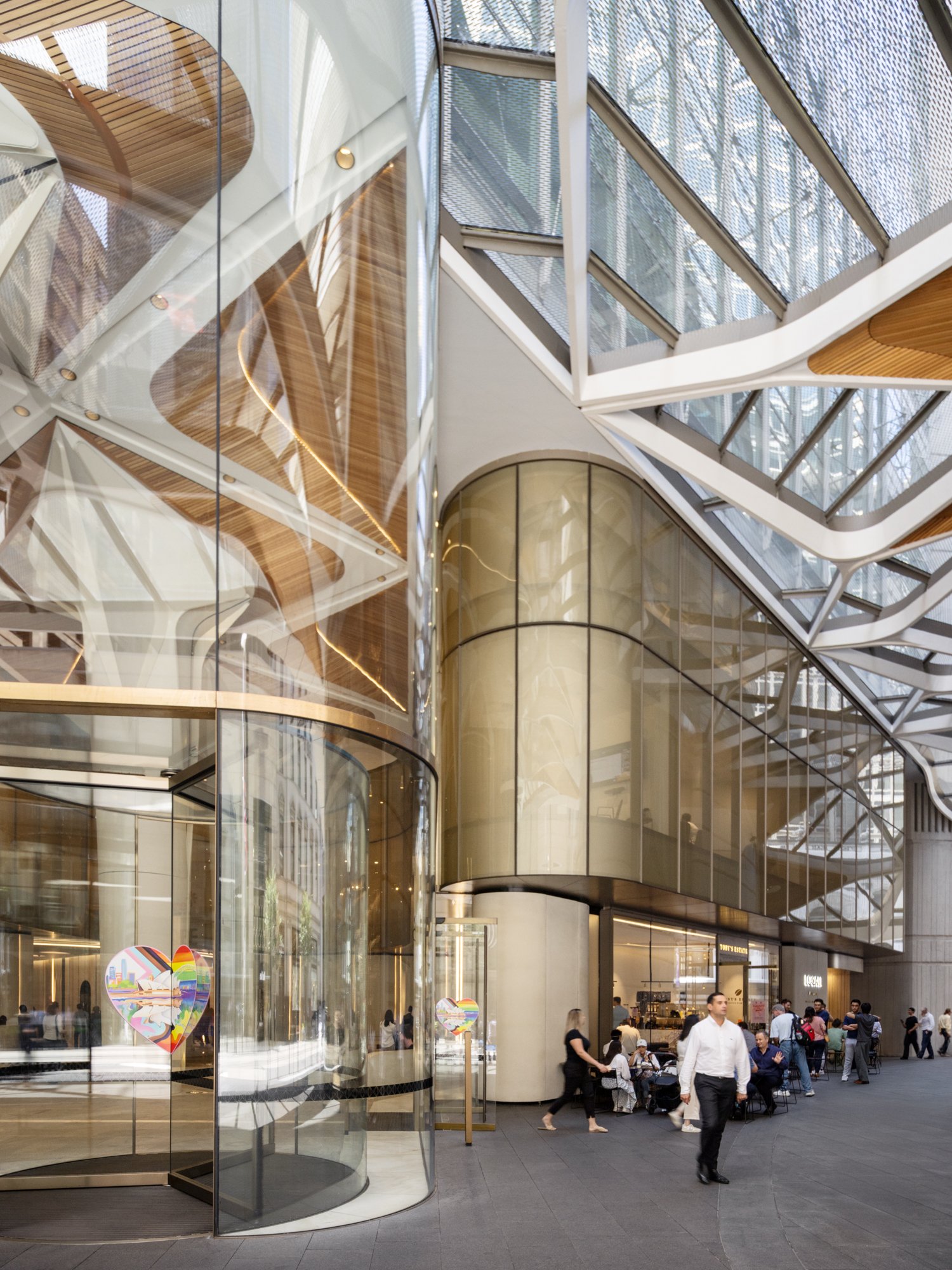 Interior view of a modern shopping mall with glass and metal architecture, people walking and sitting outside shops. Designed by fjcStudio Photo by Ruth Gold