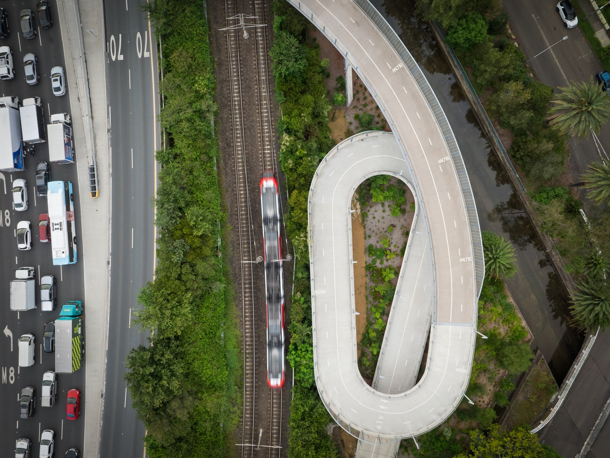 A bird's eye view of a multi-lane highway with traffic, a train passing while moving on a railway track, and a curved pedestrian bridge surrounded by green trees and small landscaped areas. Designed by Colin Polwarth Studio Photo by Ruth Gold