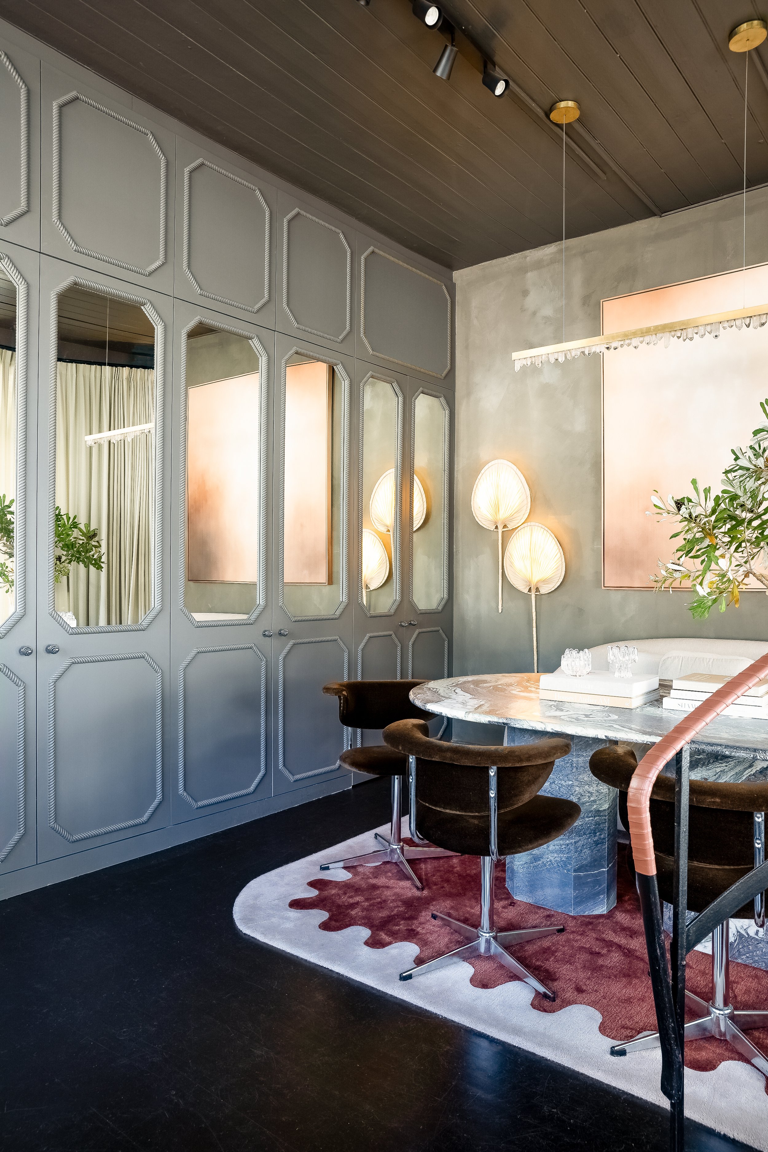 A stylish dining room featuring a marble table, black velvet chairs, decorative lighting on the wall, a patterned rug, and paneled walls with a mirror and greenery.  SMAC Studio Photo by Ruth Gold