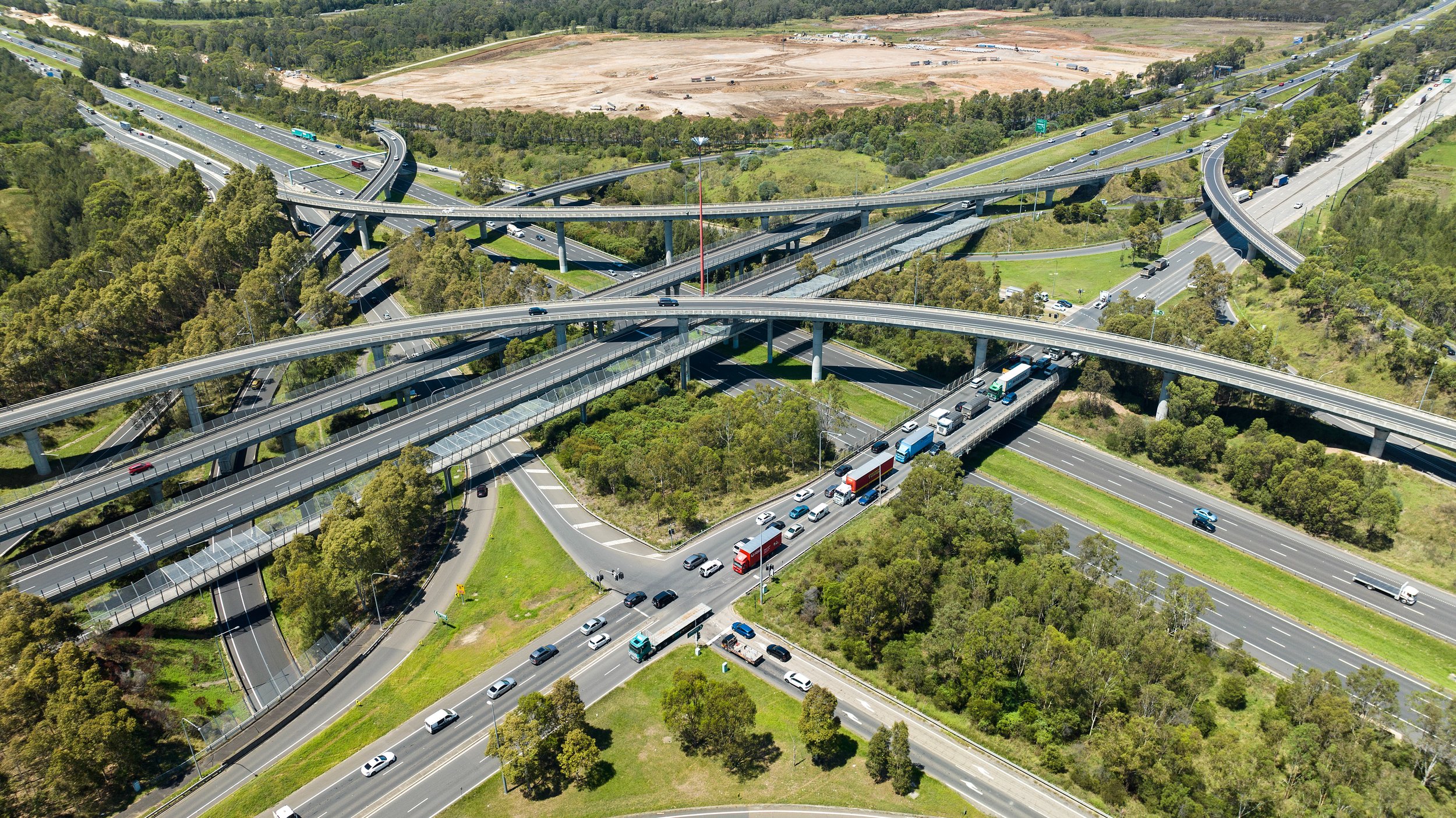 Aerial view of a multi-level highway interchange with multiple roads, overpasses, and bridges surrounded by green trees and a field in the background, with several vehicles including cars and trucks traveling in different directions.