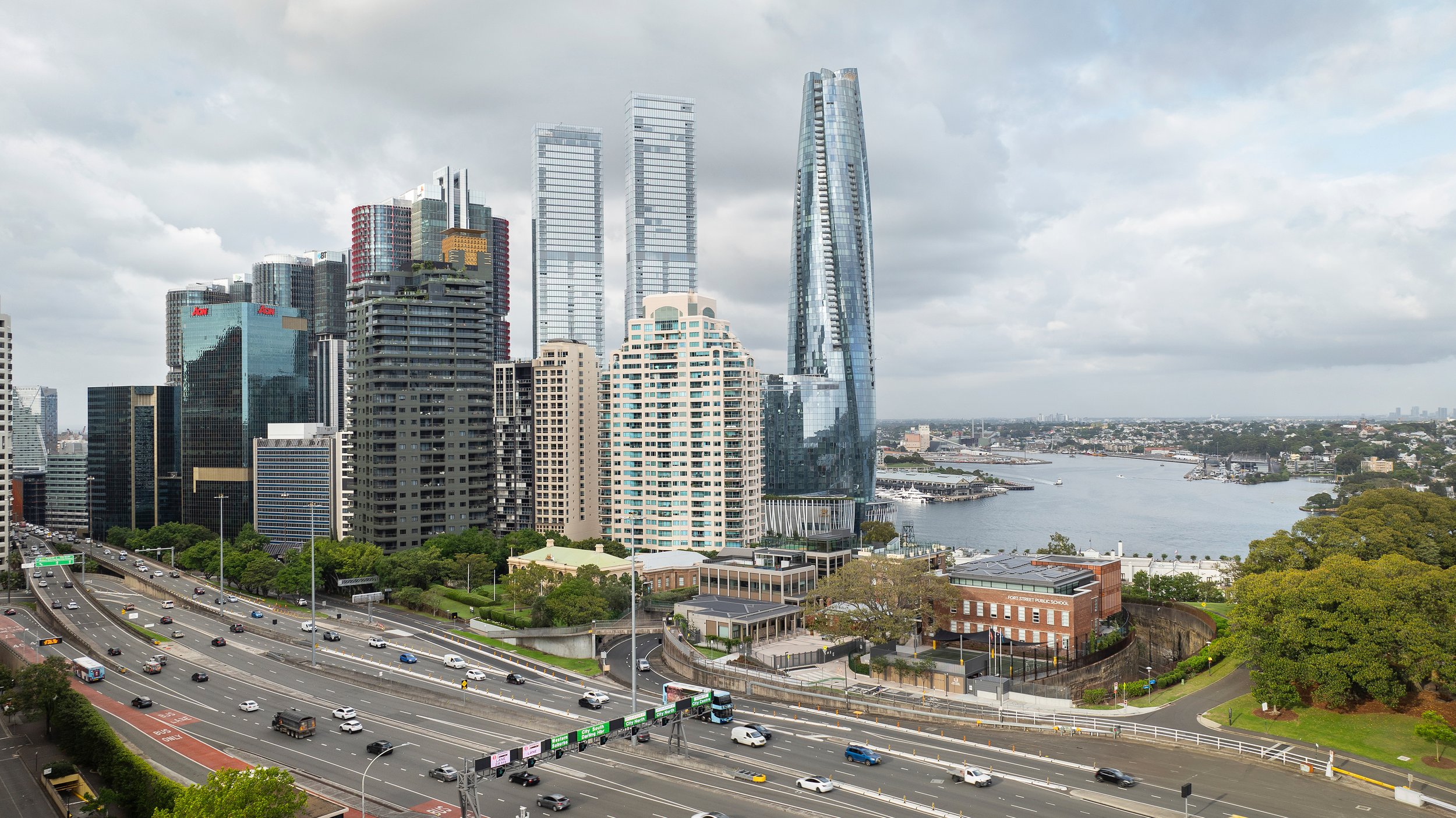 City skyline with tall office buildings and skyscrapers near a body of water, with highways and green spaces in the foreground. Designed by fjcStudio Photo by Ruth Gold