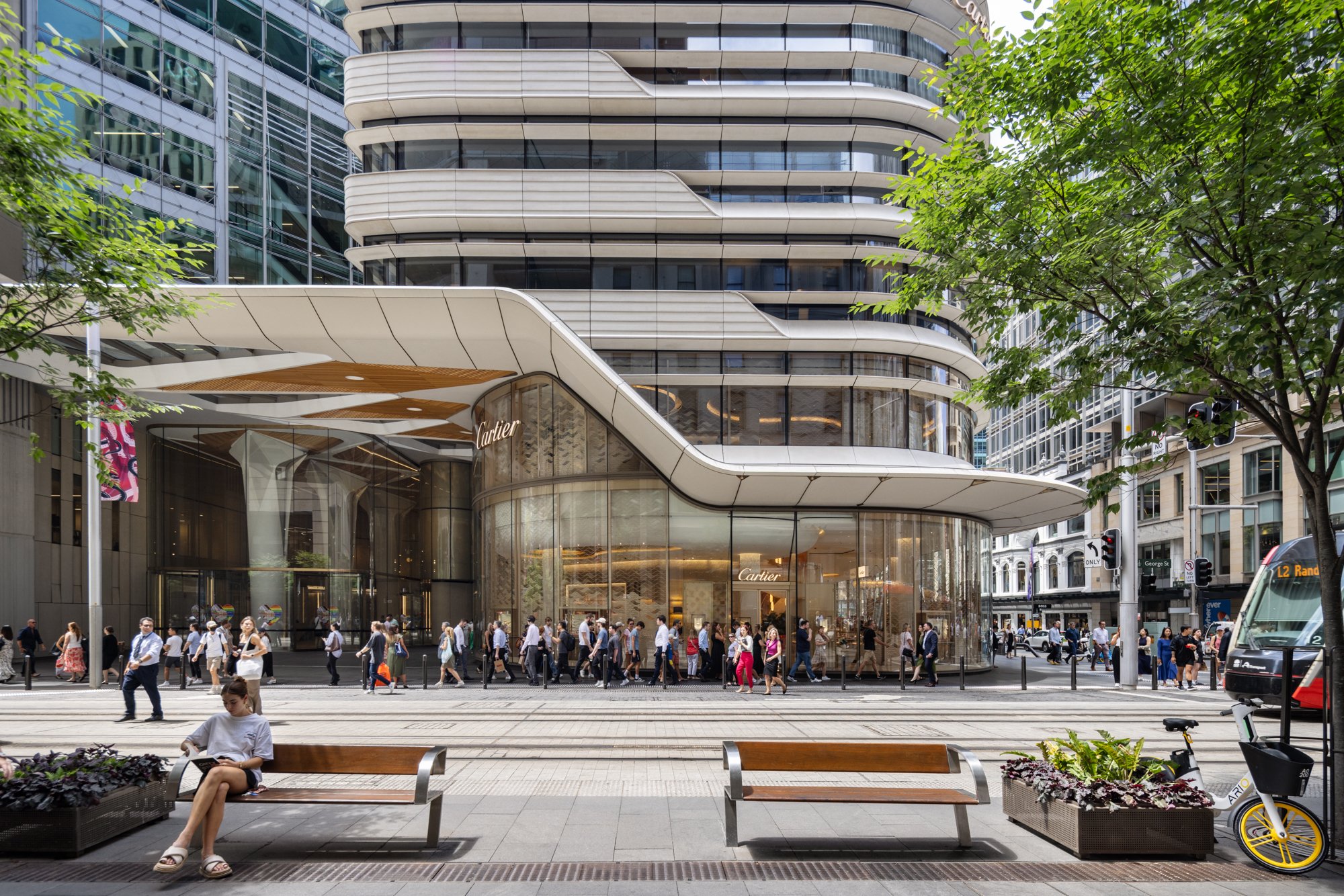 A busy city street scene featuring a modern glass and steel building with boutique storefronts, including Cartier, with pedestrians walking along the sidewalk, a person sitting on a bench reading, trees, benches, a bike, and a tram in the background.