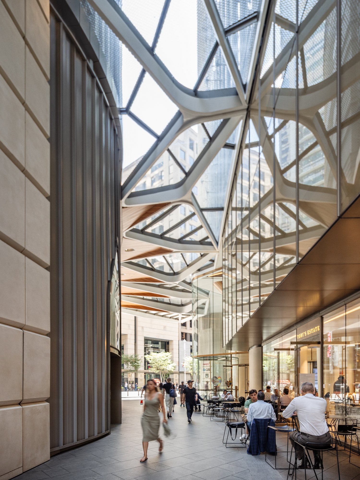 Modern indoor shopping plaza with geometric glass ceiling, outdoor seating area with several people, storefronts, and cityscape view. Designed by fjcStudio Photo by Ruth Gold