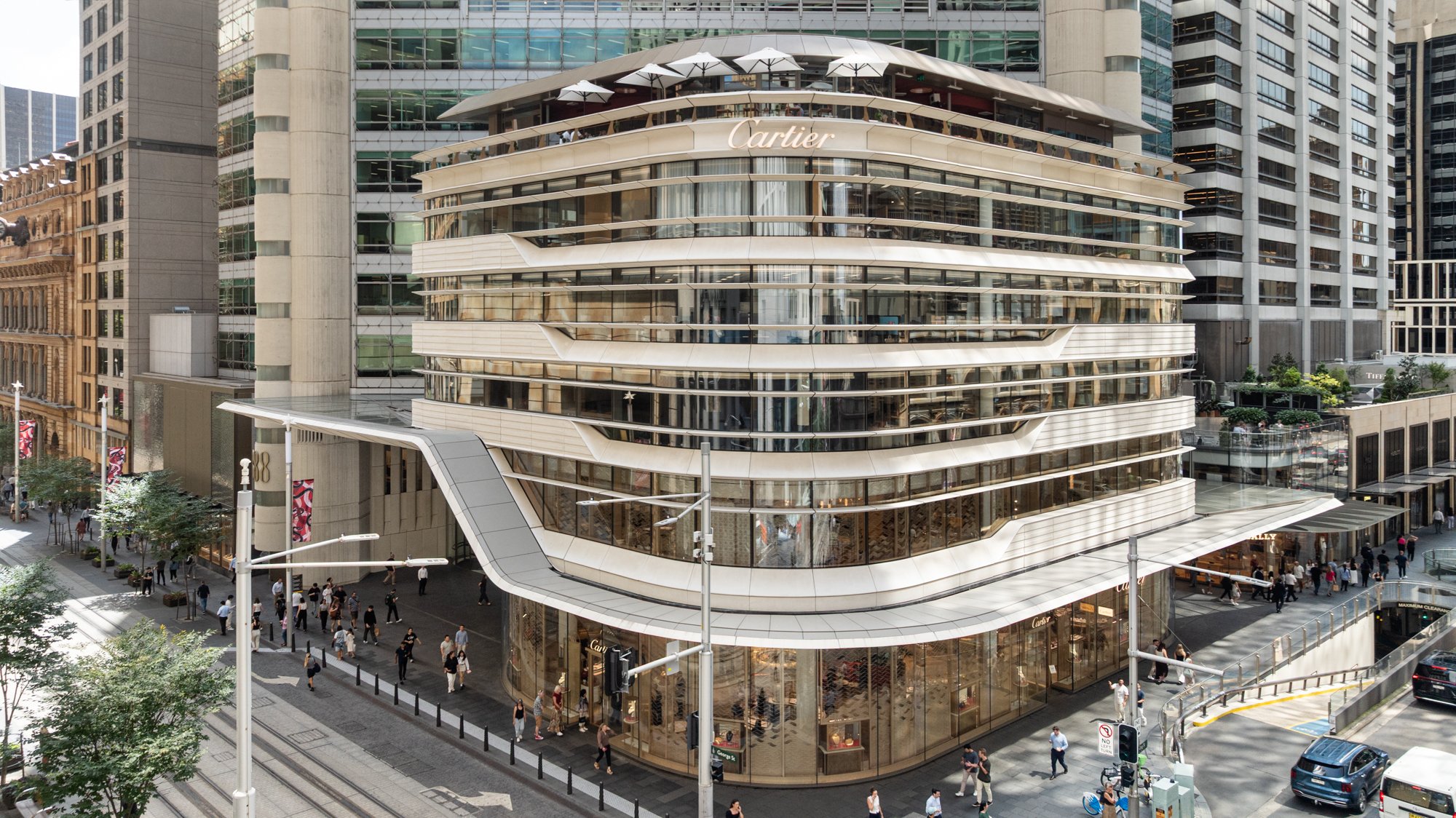 A modern multi-story shopping mall with the Cartier store on the upper levels, busy street with pedestrians, cars, and trees in an urban setting. Designed by fjcStudio Photo by Ruth Gold