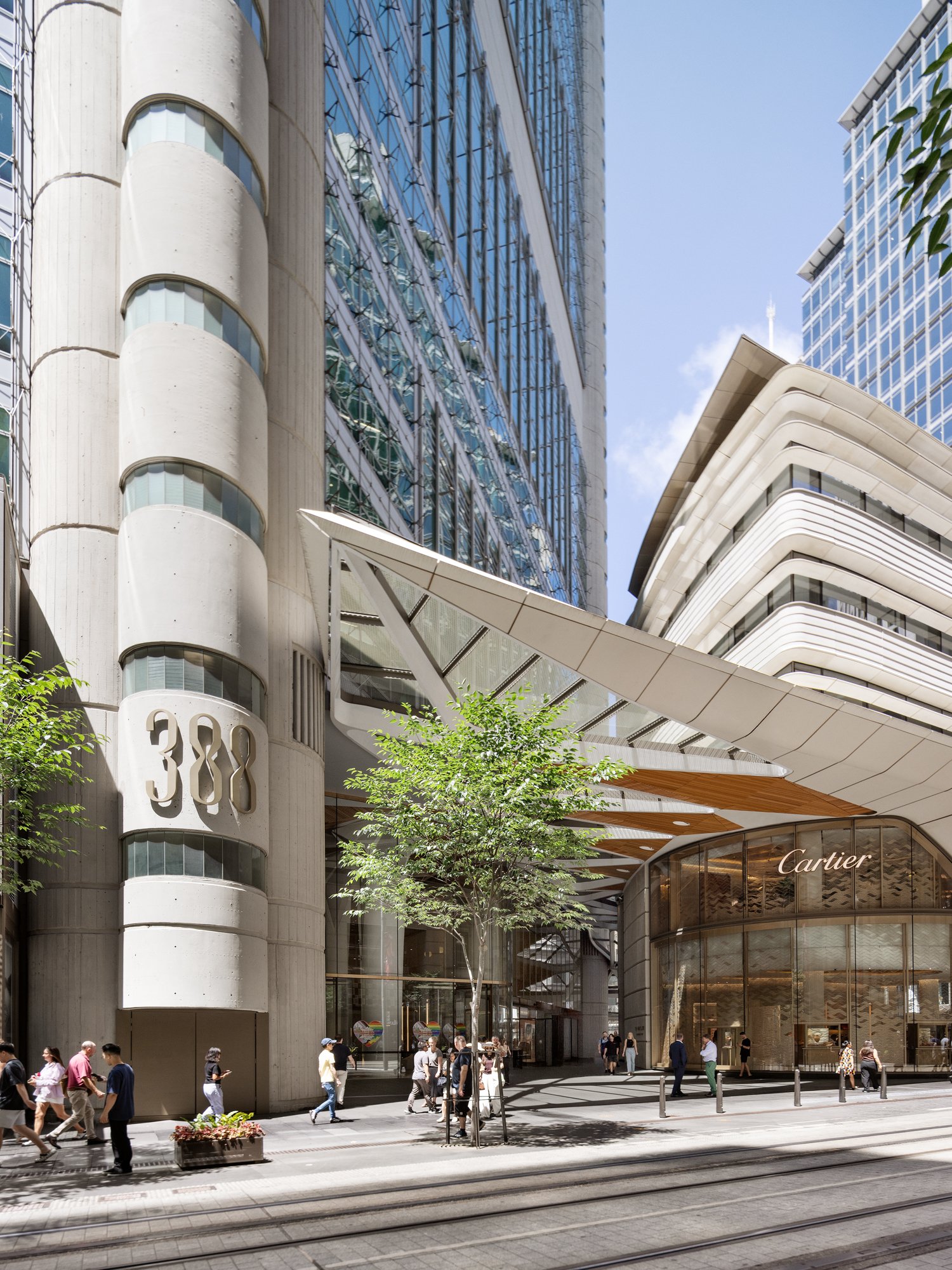 Modern city street scene with tall glass buildings, a Cartier store, pedestrians walking, trees, and tram tracks. Designed by fjcStudio Photo by Ruth Gold