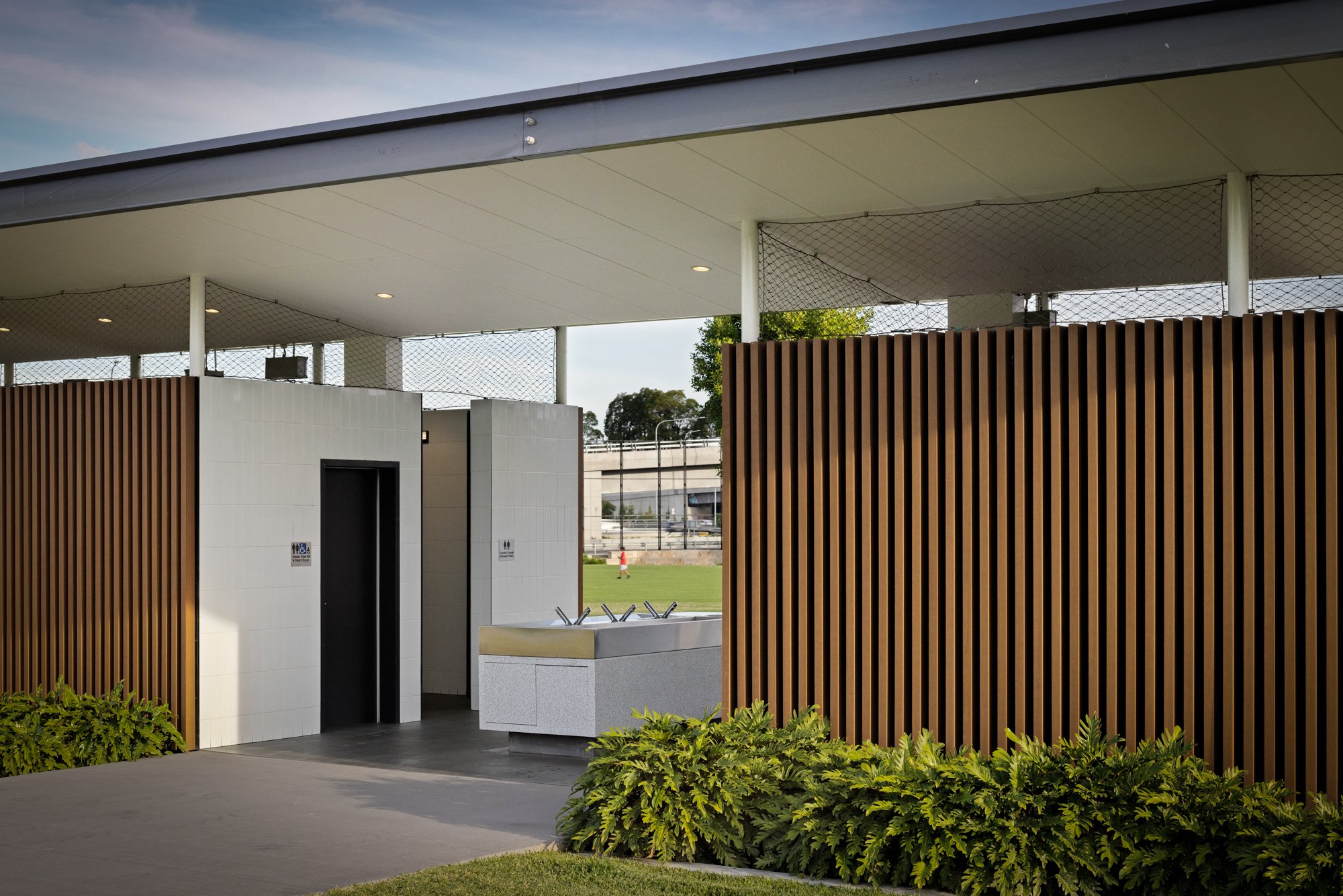 Public restroom facilities with a white tiled wall, black door, water fountain, and wooden slat privacy wall, outdoor setting with grass and trees. Designed by Colin Polwarth Studio Photo by Ruth Gold