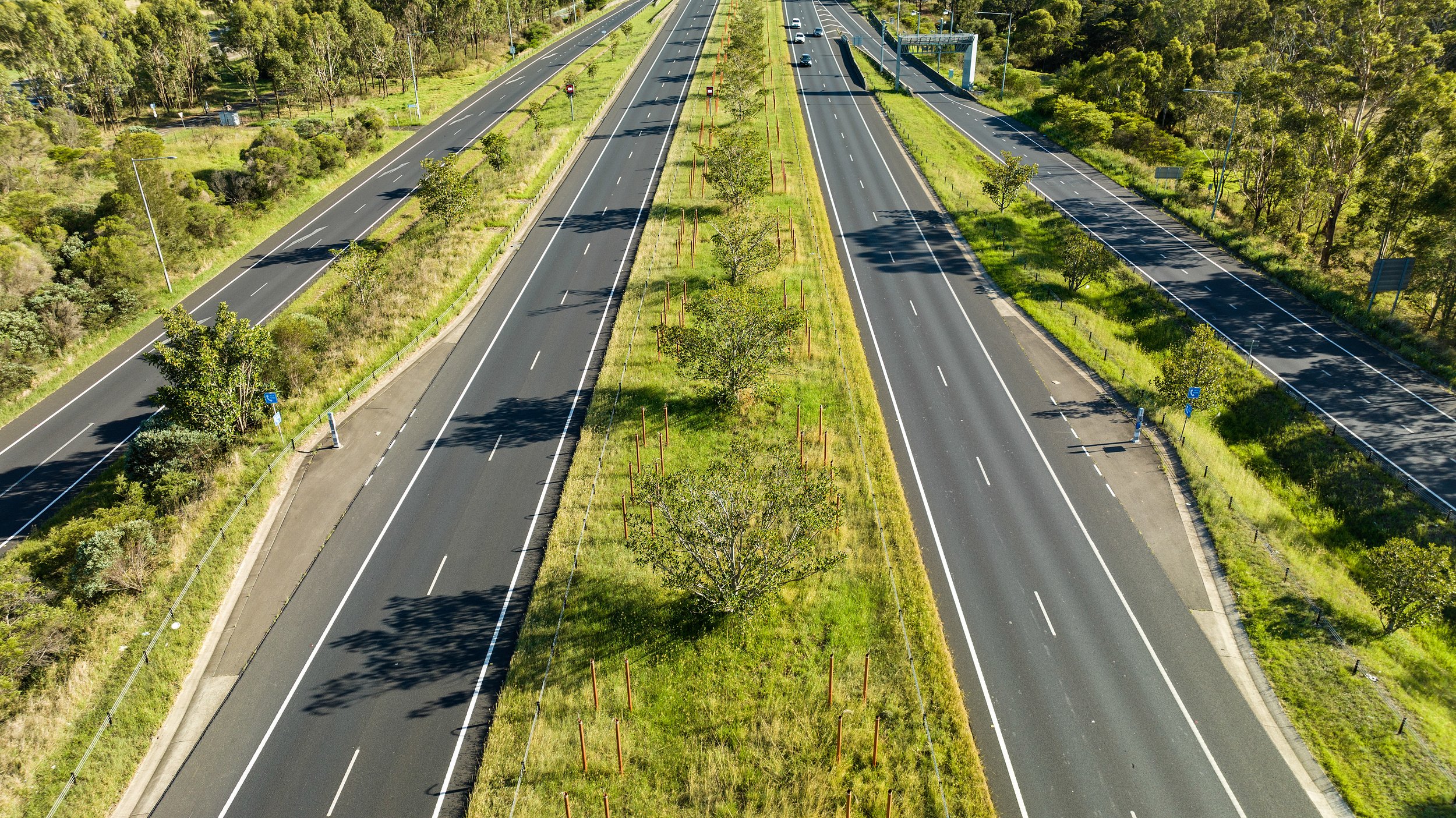 An aerial view of a divided highway with three lanes in each direction, surrounded by green trees and grass, with some trees planted along the median. Designed by Colin Polwarth Studio Photo by Ruth Gold