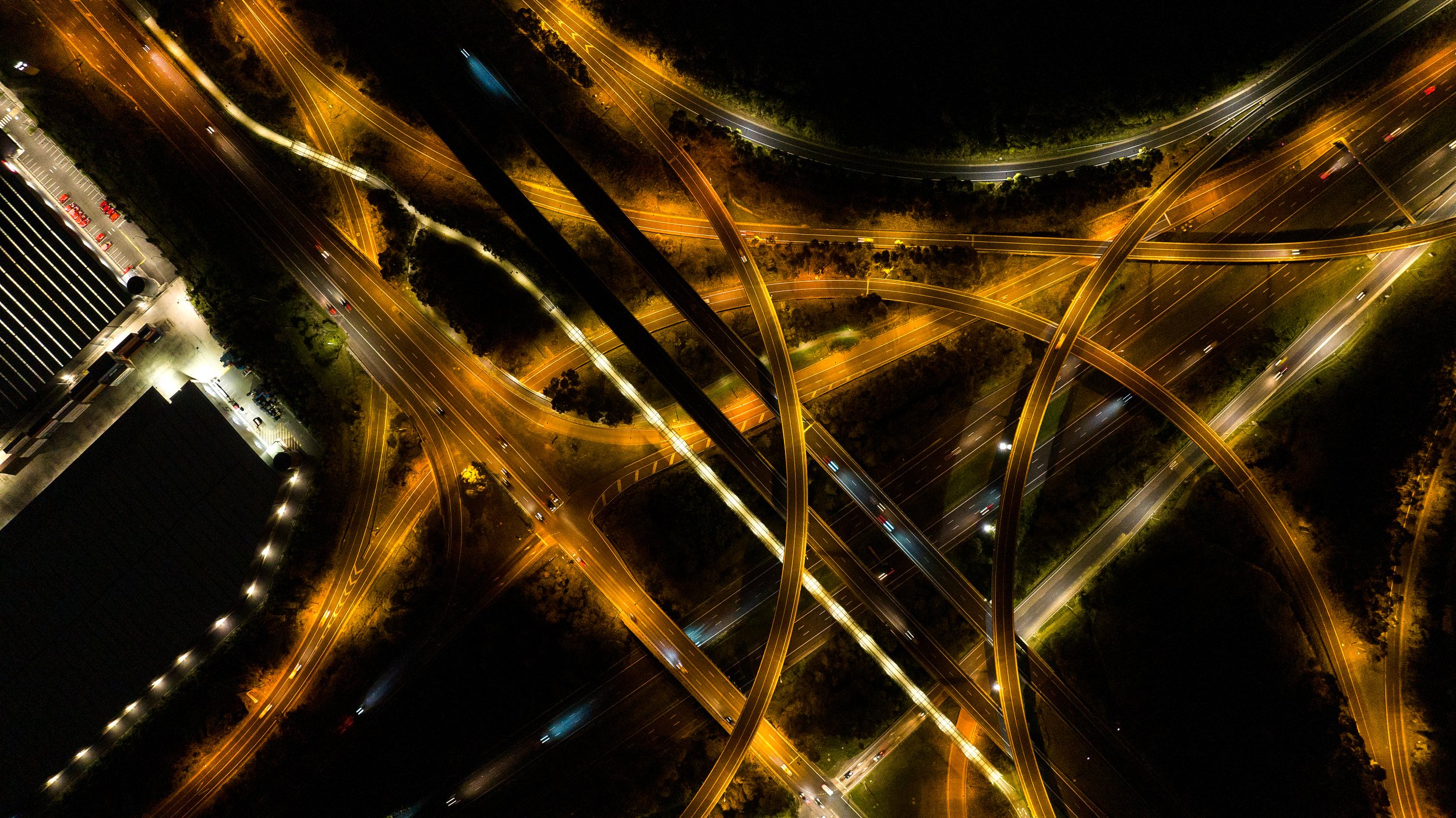 Night aerial view of a complex highway interchange with multiple overpasses and roads illuminated by streetlights. Designed by Colin Polwarth Studio Photo by Ruth Gold