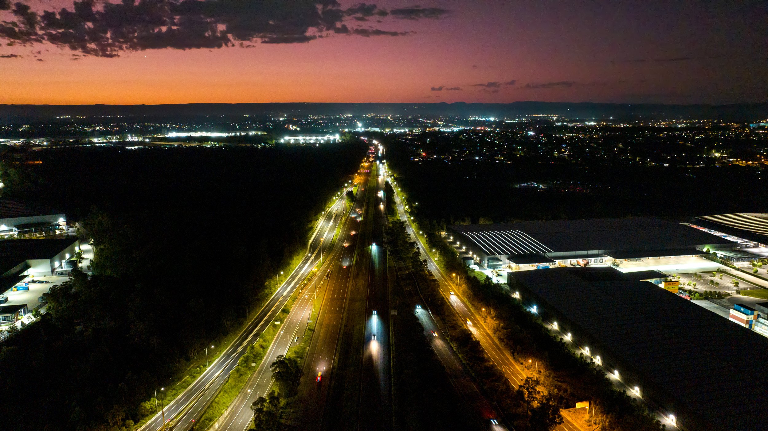 An aerial view of a highway at dusk with city lights in the background and a colorful sunset in the sky. Designed by Colin Polwarth Studio Photo by Ruth Gold