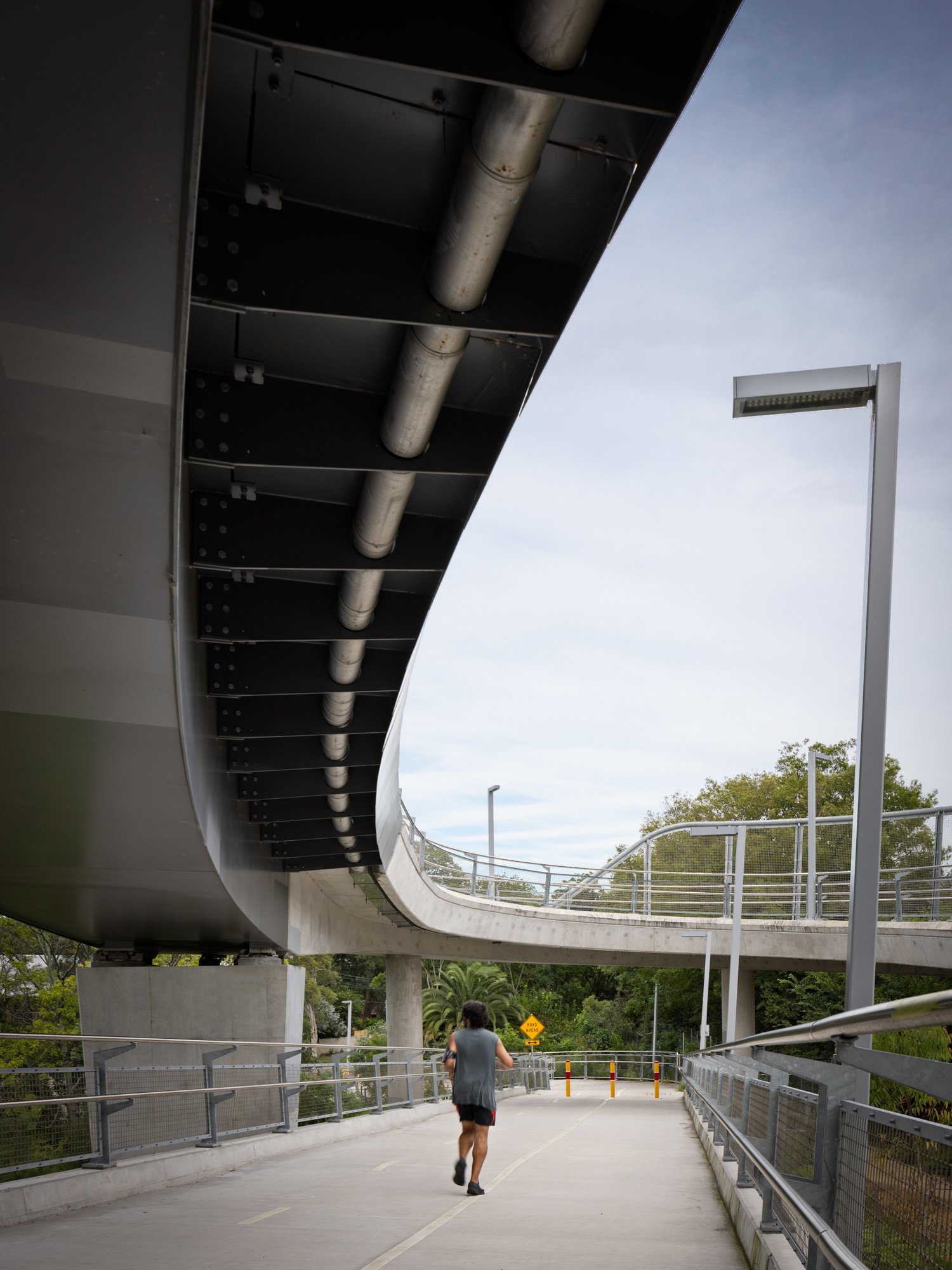 A person jogging underneath a modern, elevated concrete and metal pedestrian bridge with a large metal pipe underneath and a cloudy sky above. Designed by Colin Polwarth Studio Photo by Ruth Gold