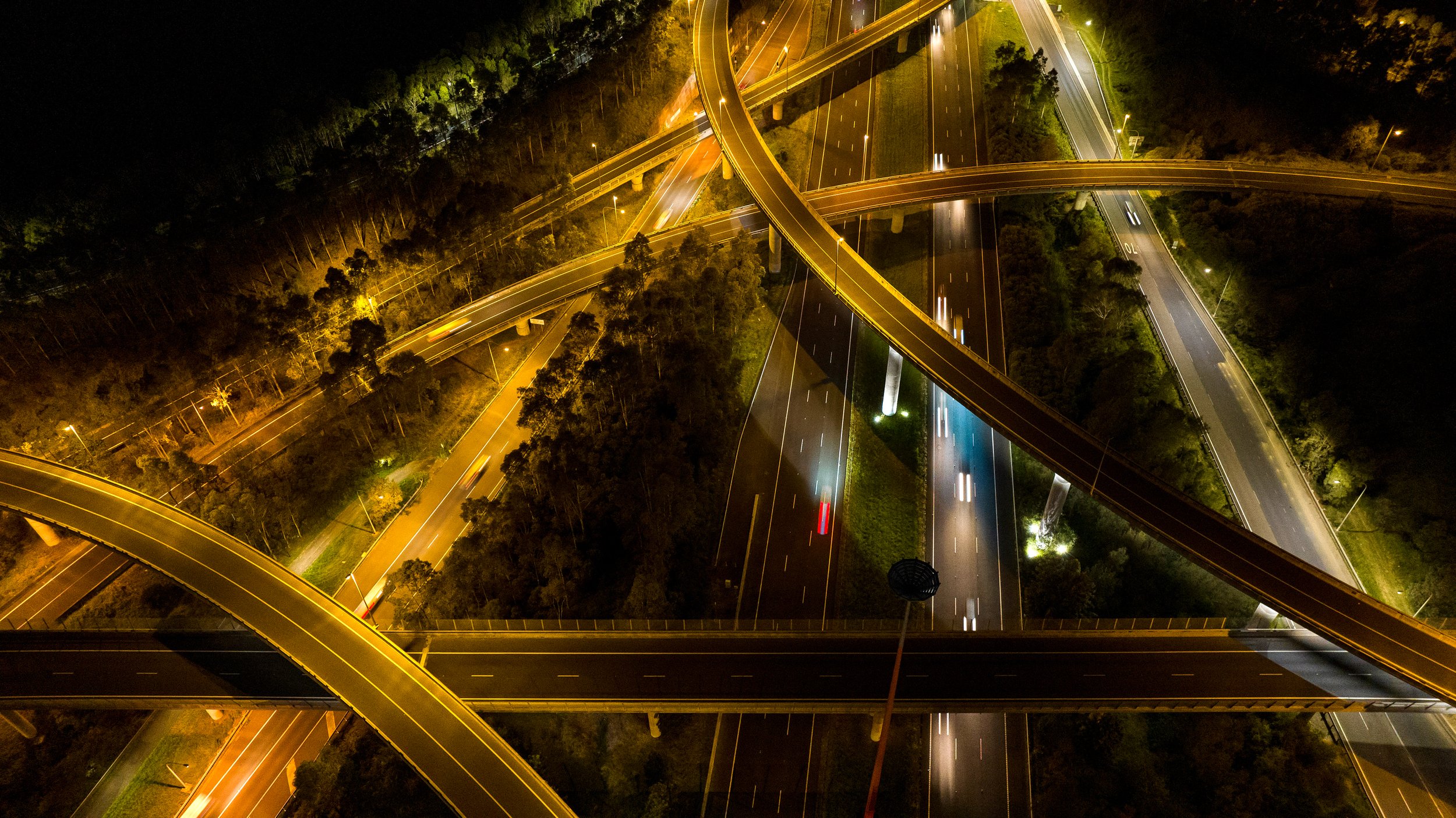 Night aerial view of a multi-level highway interchange illuminated with streetlights and car lights, surrounded by trees and dark landscape. Designed by Colin Polwarth Studio Photo by Ruth Gold