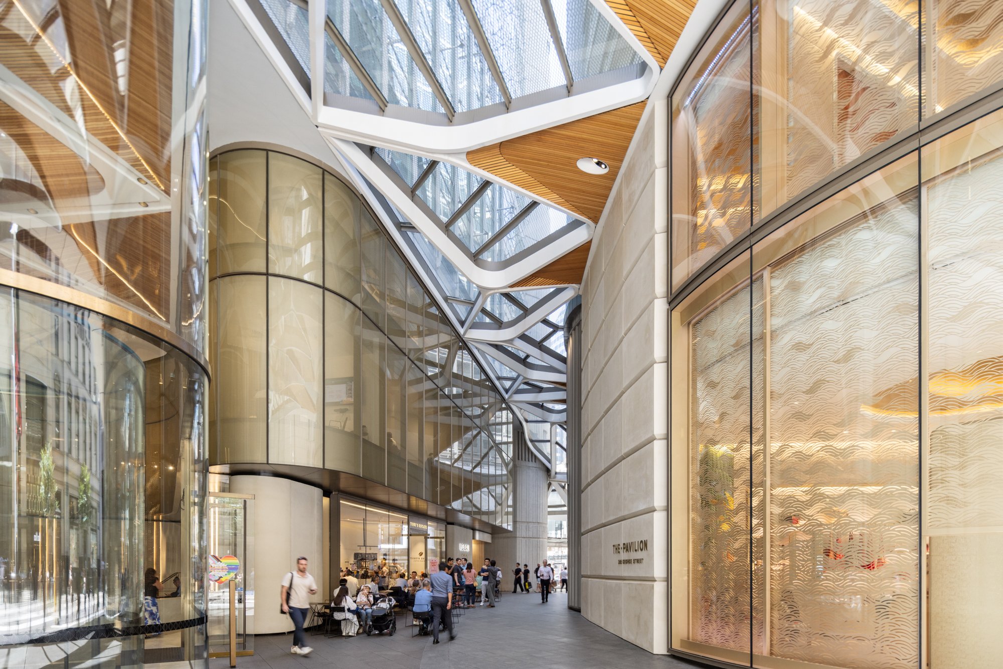 Interior view of a modern shopping mall with curved glass walls, contemporary architectural design, and shoppers walking and sitting at tables. Designed by fjcStudio Photo by Ruth Gold
