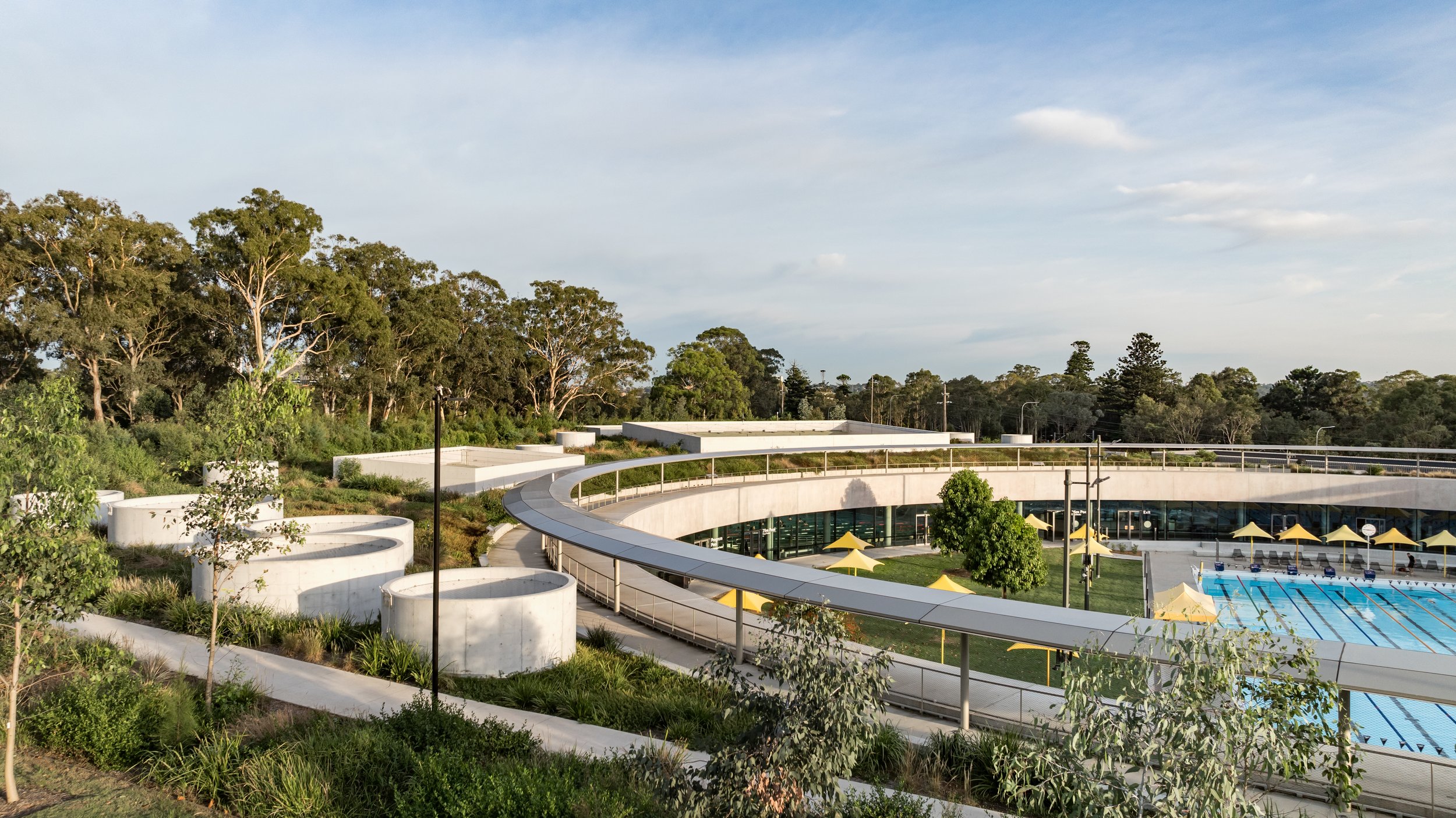 A modern outdoor swimming pool area with a curved walkway, surrounded by green trees and shrubbery. The pool has yellow umbrellas along the edge, and a backdrop of trees and a blue sky with some clouds. Designed by McGregor Coxall Photo by Ruth Gold