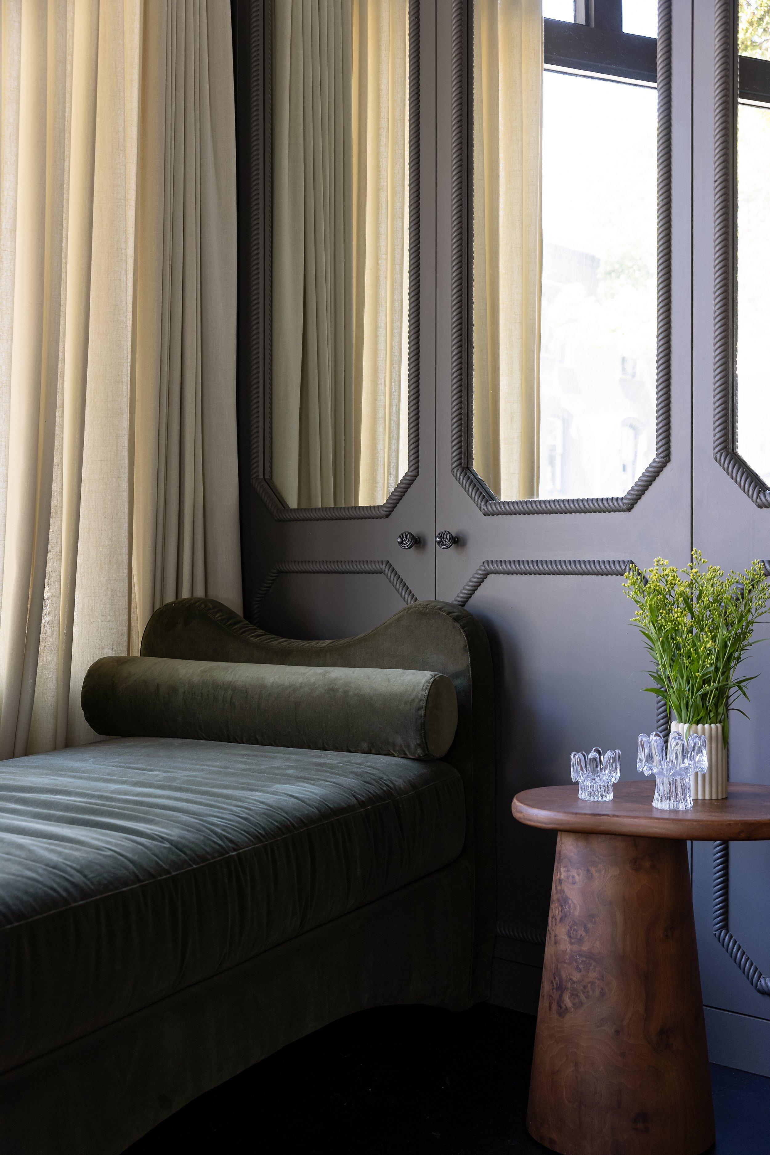 A dark green velvet chaise lounge next to a wooden side table with glass candle holders and a vase of yellow flowers, near a window with beige curtains and gray decorative doors. SMAC Studio Photo by Ruth Gold