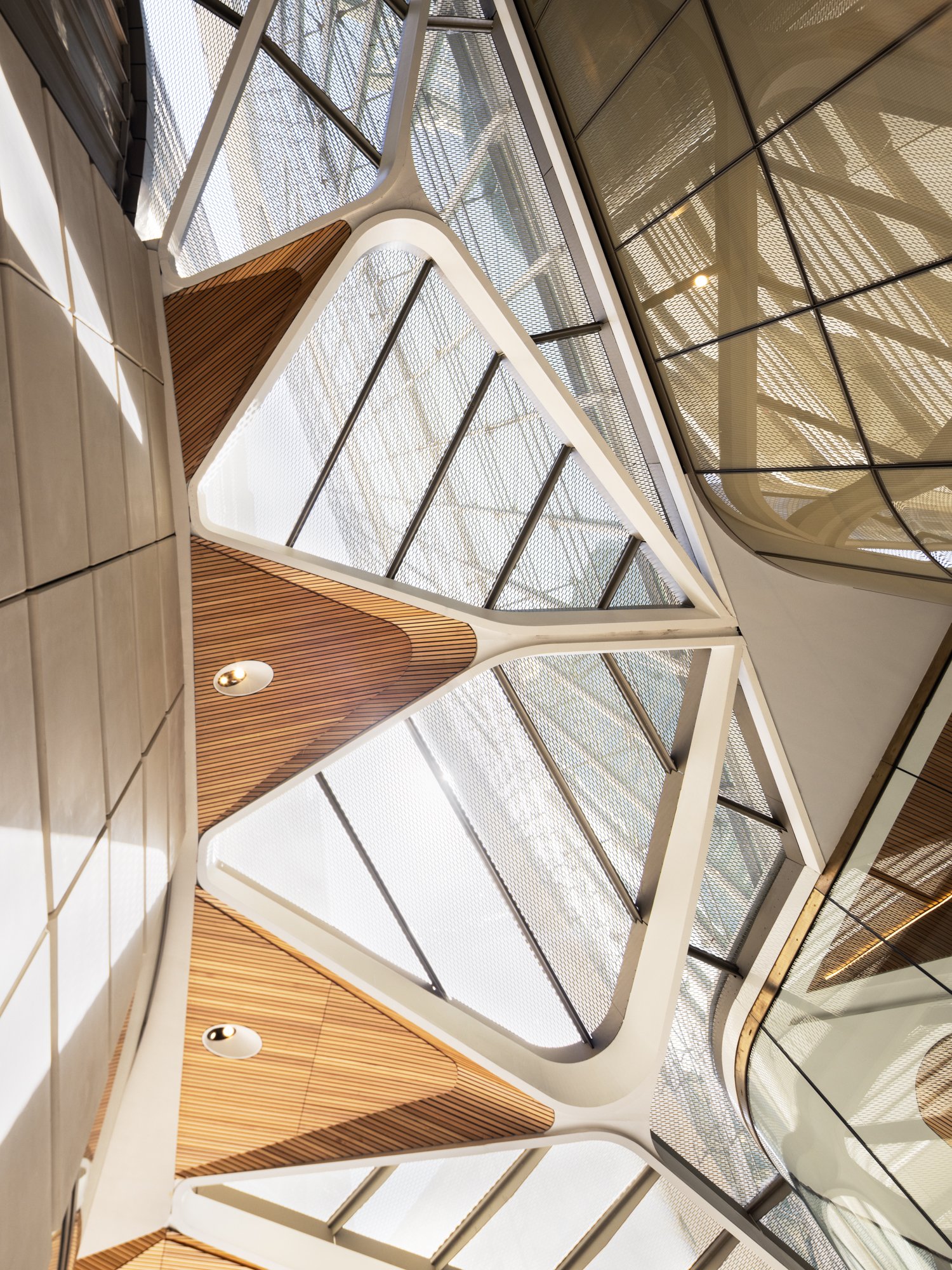 View of a modern architectural interior ceiling with geometric shapes, wooden panels, glass, and metal mesh structures, emphasizing natural light and contemporary design. Designed by fjcStudio Photo by Ruth Gold