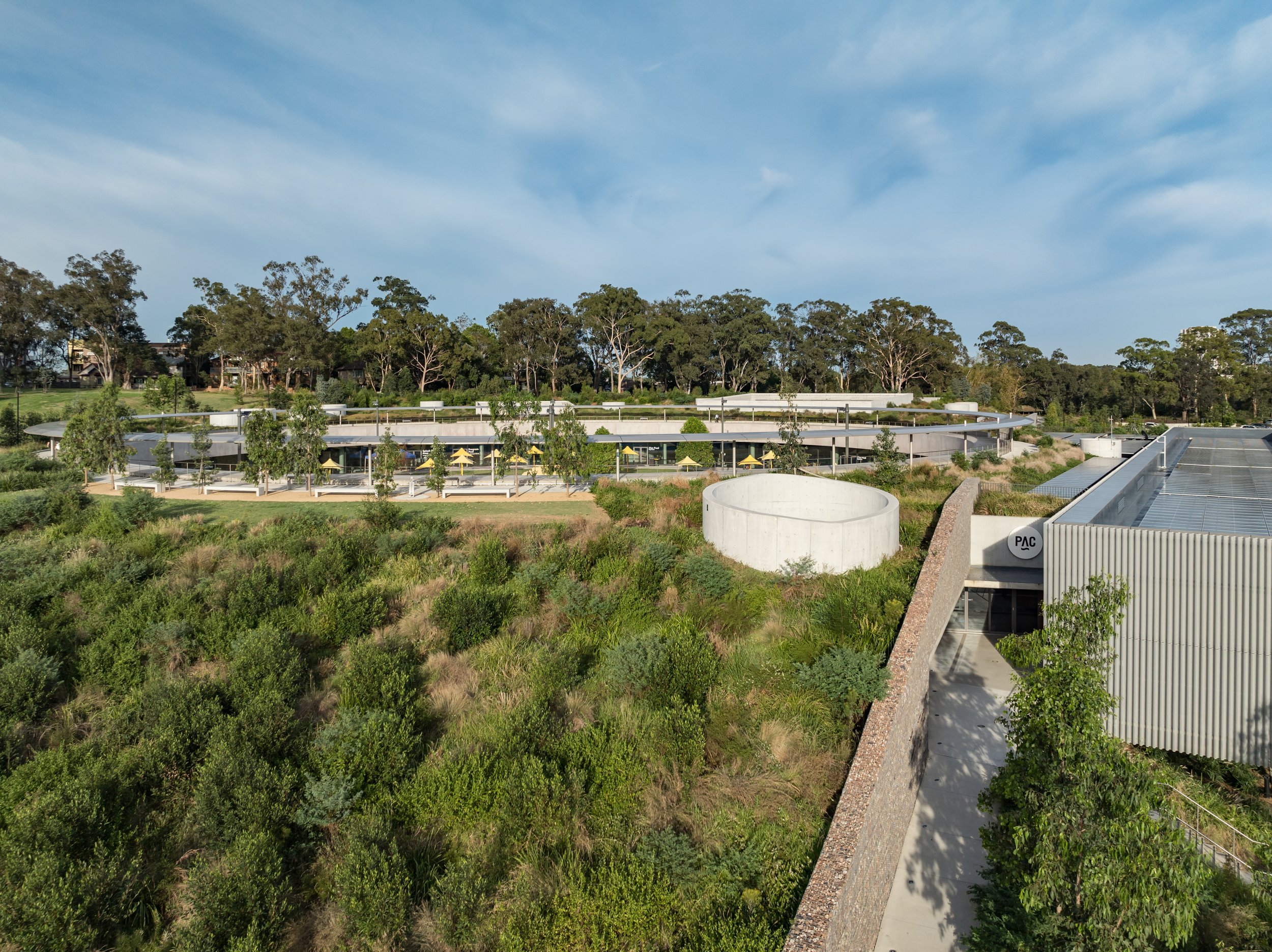 A modern building complex with solar panels on the roof, surrounded by lush greenery and a strip of trees in the background, under a partly cloudy sky. Designed by McGregor Coxall Photo by Ruth Gold