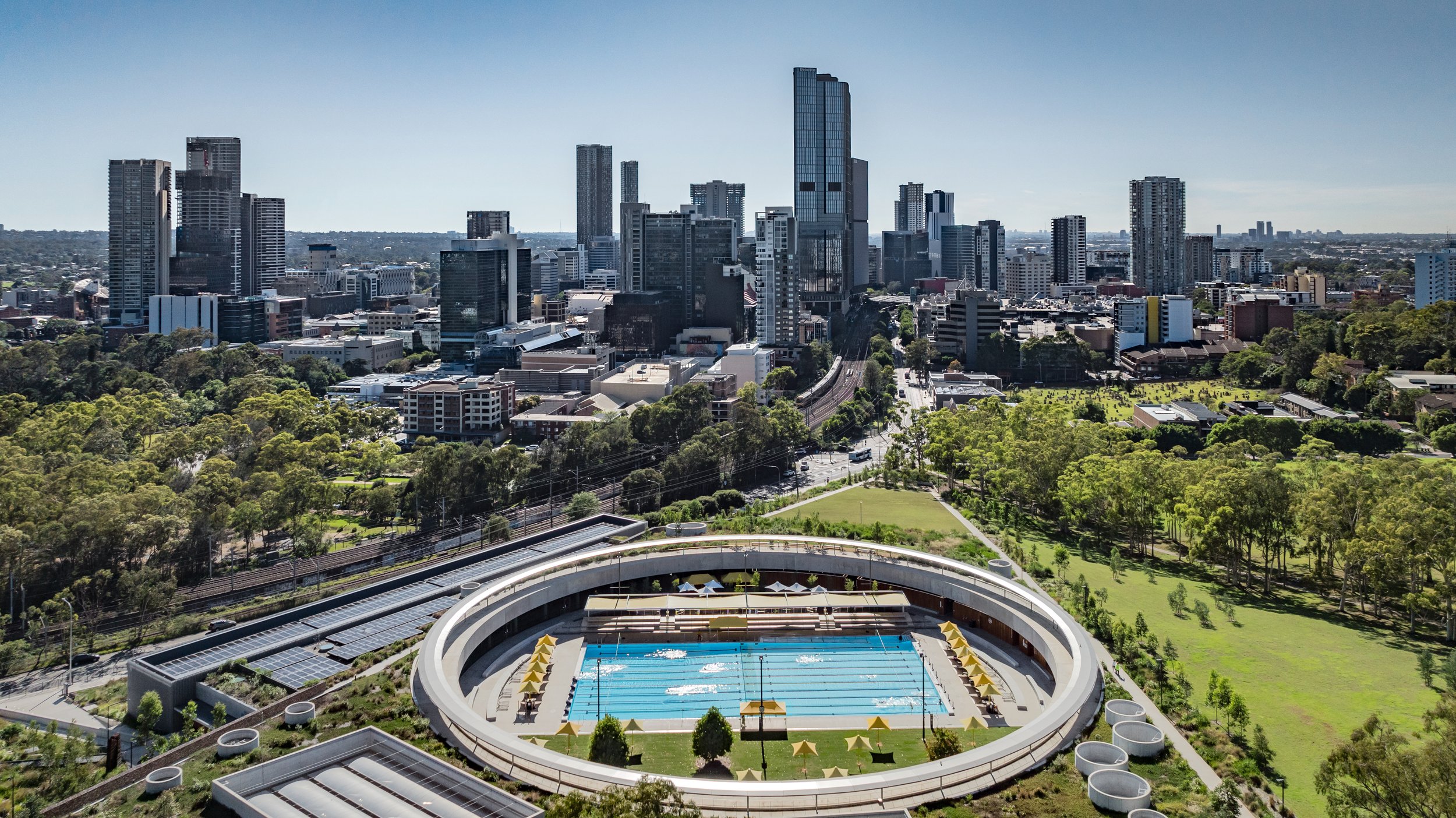 Aerial view of a city skyline with tall skyscrapers, a large outdoor swimming pool, and green park areas. Designed by McGregor Coxall Photo by Ruth Gold