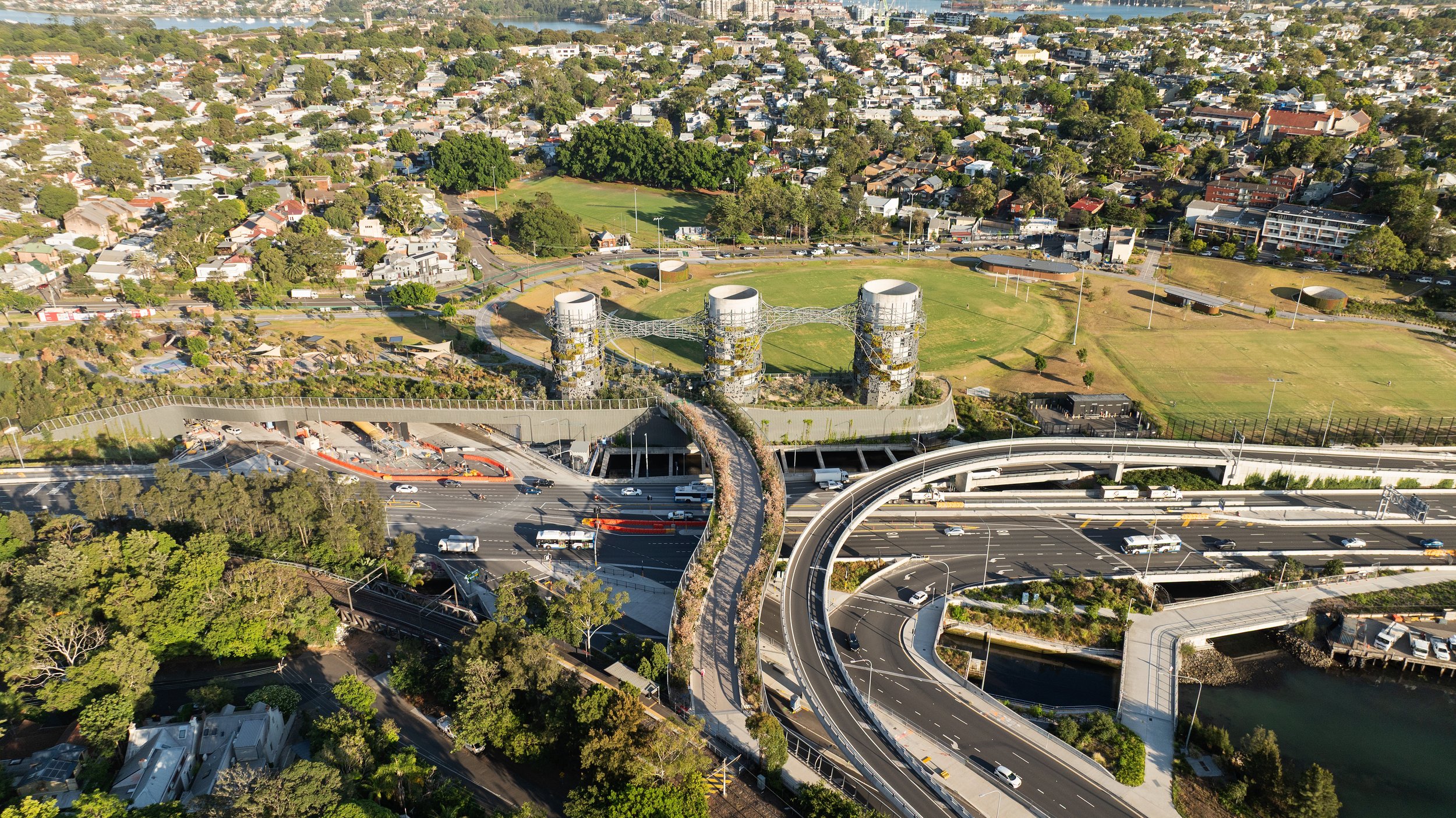 Aerial view of a cityscape featuring highways, a park with three cooling towers, green residential neighborhoods, and a river. Designed by McGregor Coxall Photo by Ruth Gold