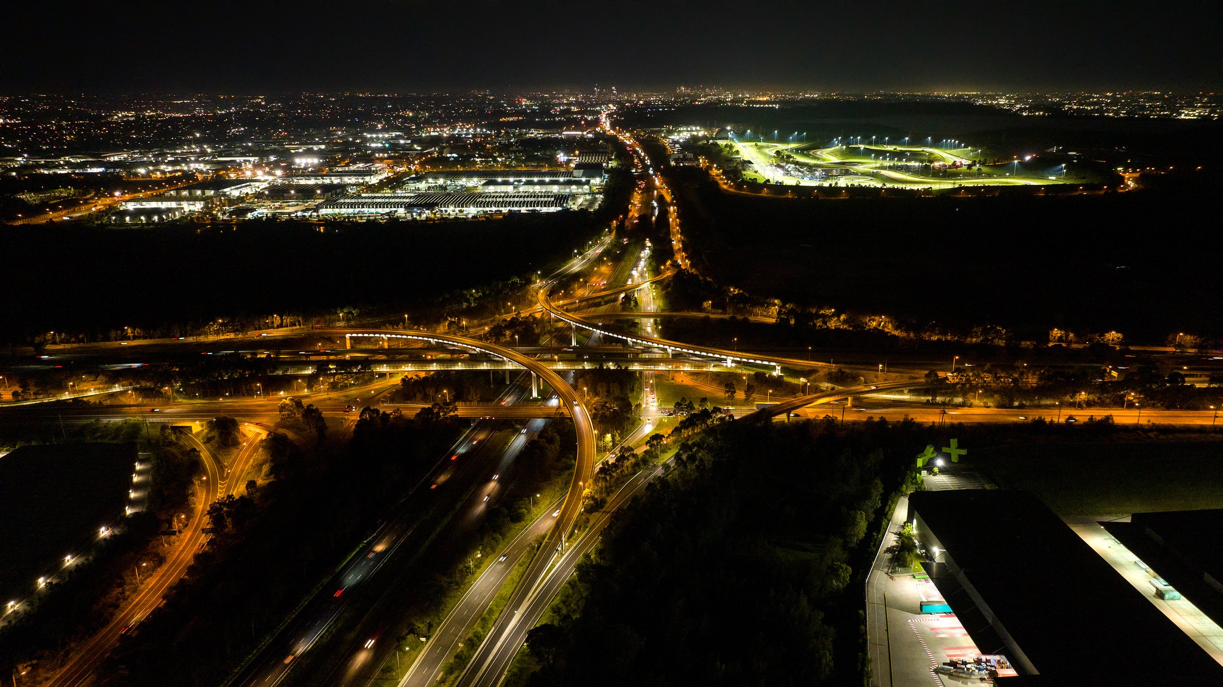Nighttime aerial view of a city with illuminated roads, highways, and buildings, with a large green pharmacy cross sign visible in the lower right corner. Designed by Colin Polwarth Studio Photo by Ruth Gold