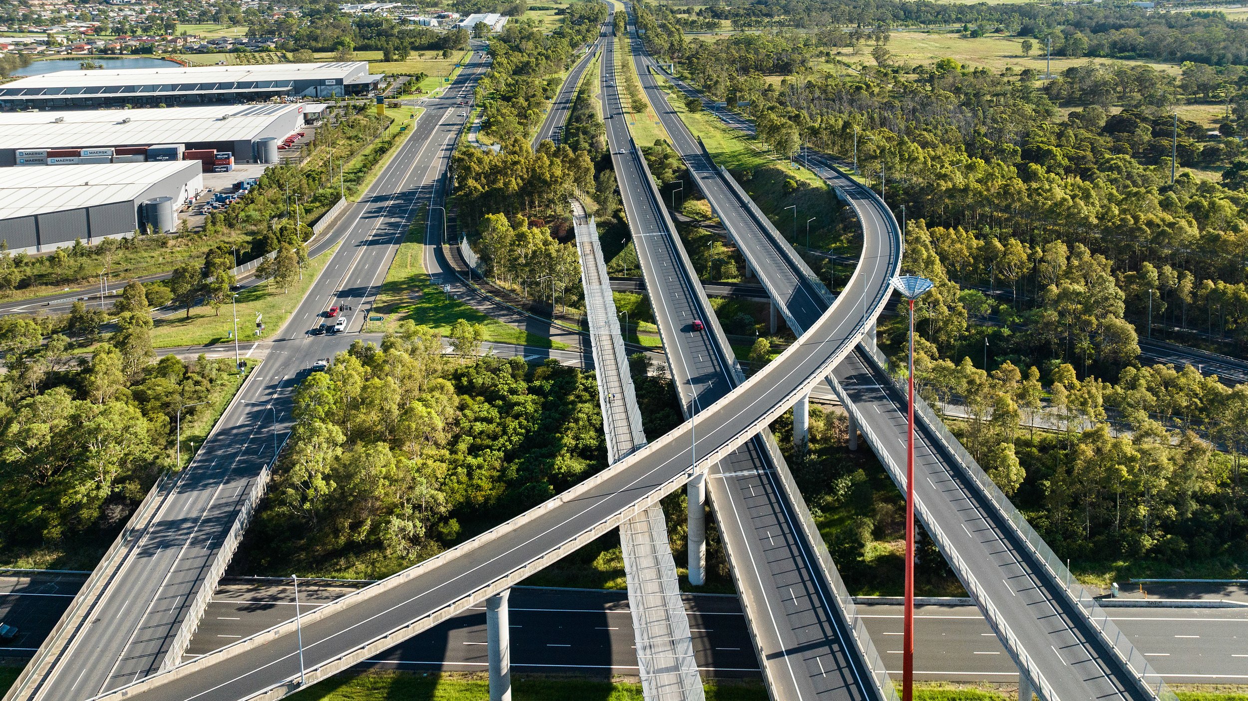 Aerial view of multiple intersecting highway overpasses and roads surrounded by green trees and industrial warehouse buildings. Designed by Colin Polwarth Studio Photo by Ruth Gold