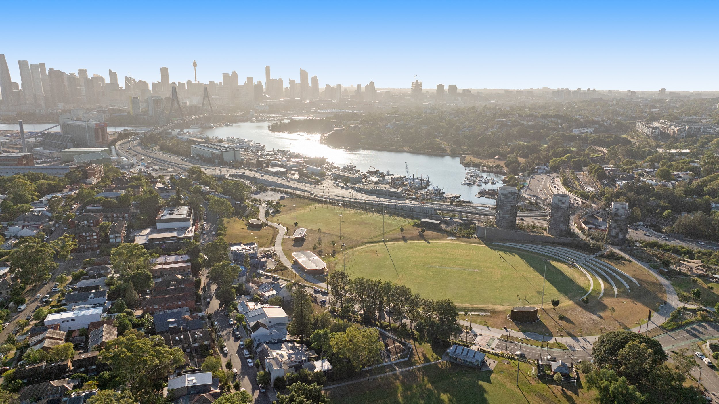 Aerial view of a city with a river, parks, residential and commercial buildings, and a bridge connecting different parts of the city, with sunny weather and clear skies. Designed by McGregor Coxall Photo by Ruth Gold