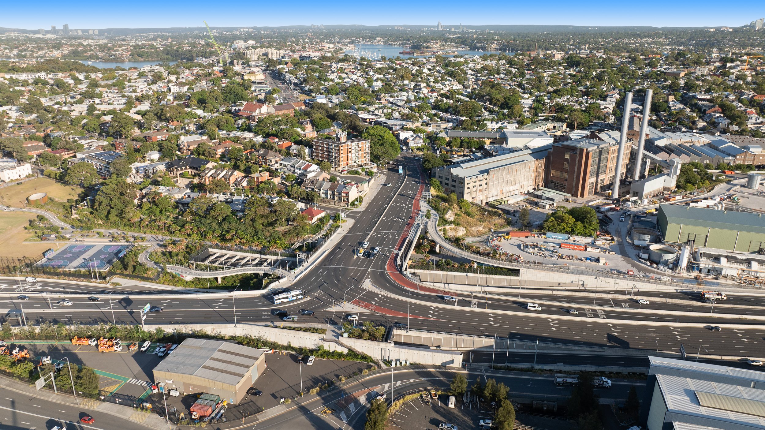 An aerial view of a cityscape featuring highways, residential buildings, industrial facilities, a sports court, and a water body in the background. Designed by McGregor Coxall Photo by Ruth Gold