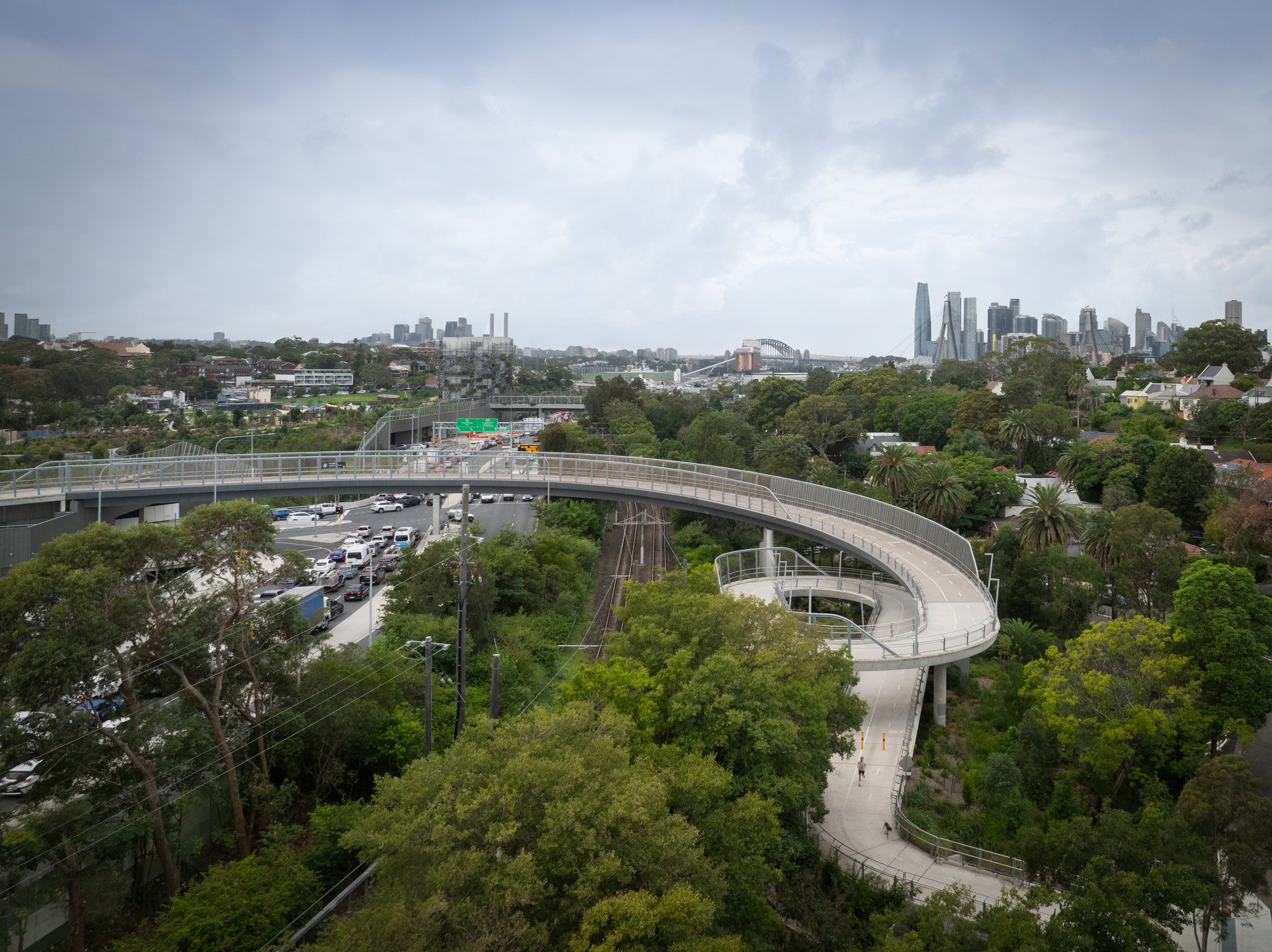 Cityscape with a curved elevated pedestrian walkway above lush green trees, traffic on the street below, and a skyline with skyscrapers and clouds in the background. Designed by Colin Polwarth Studio Photo by Ruth Gold