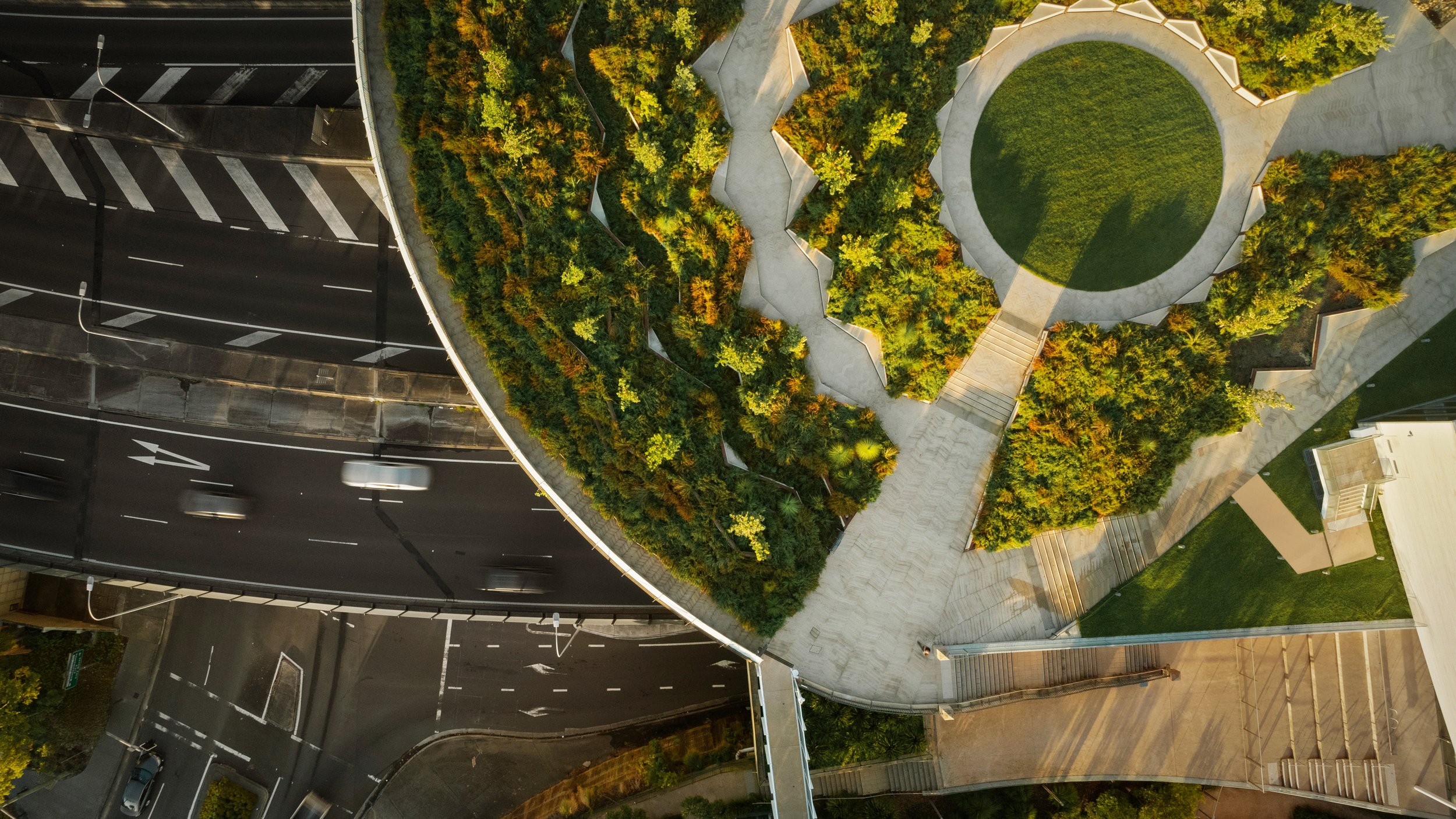 An aerial view of a city scene showing a section of a road with moving cars, a green park with walking paths, and surrounding buildings. Designed by McGregor Coxall Photo by Ruth Gold