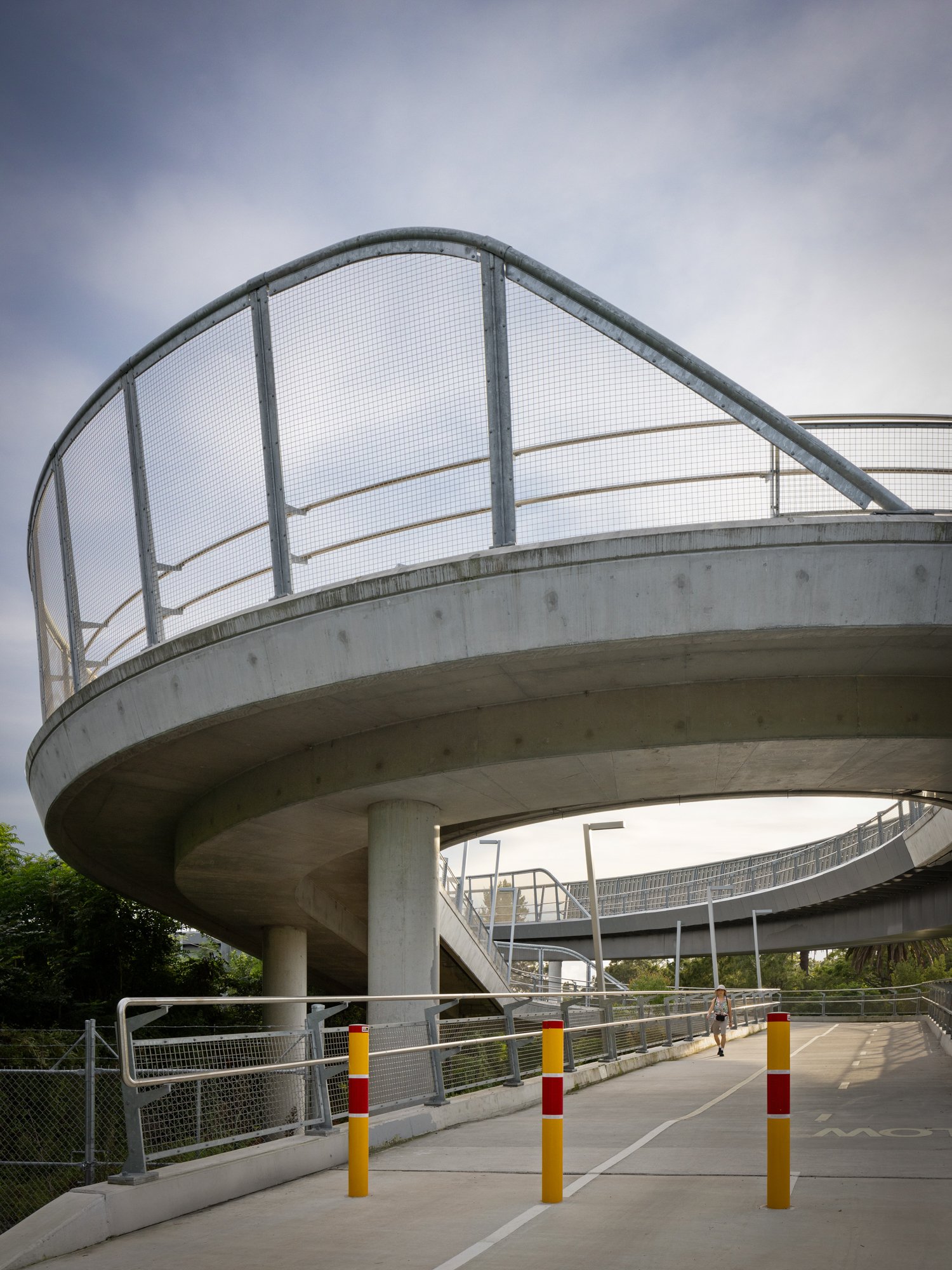 A concrete pedestrian bridge with a landscaped ramp, metal fencing, and a lone person walking underneath, with a cloudy sky overhead. Designed by Colin Polwarth Studio Photo by Ruth Gold