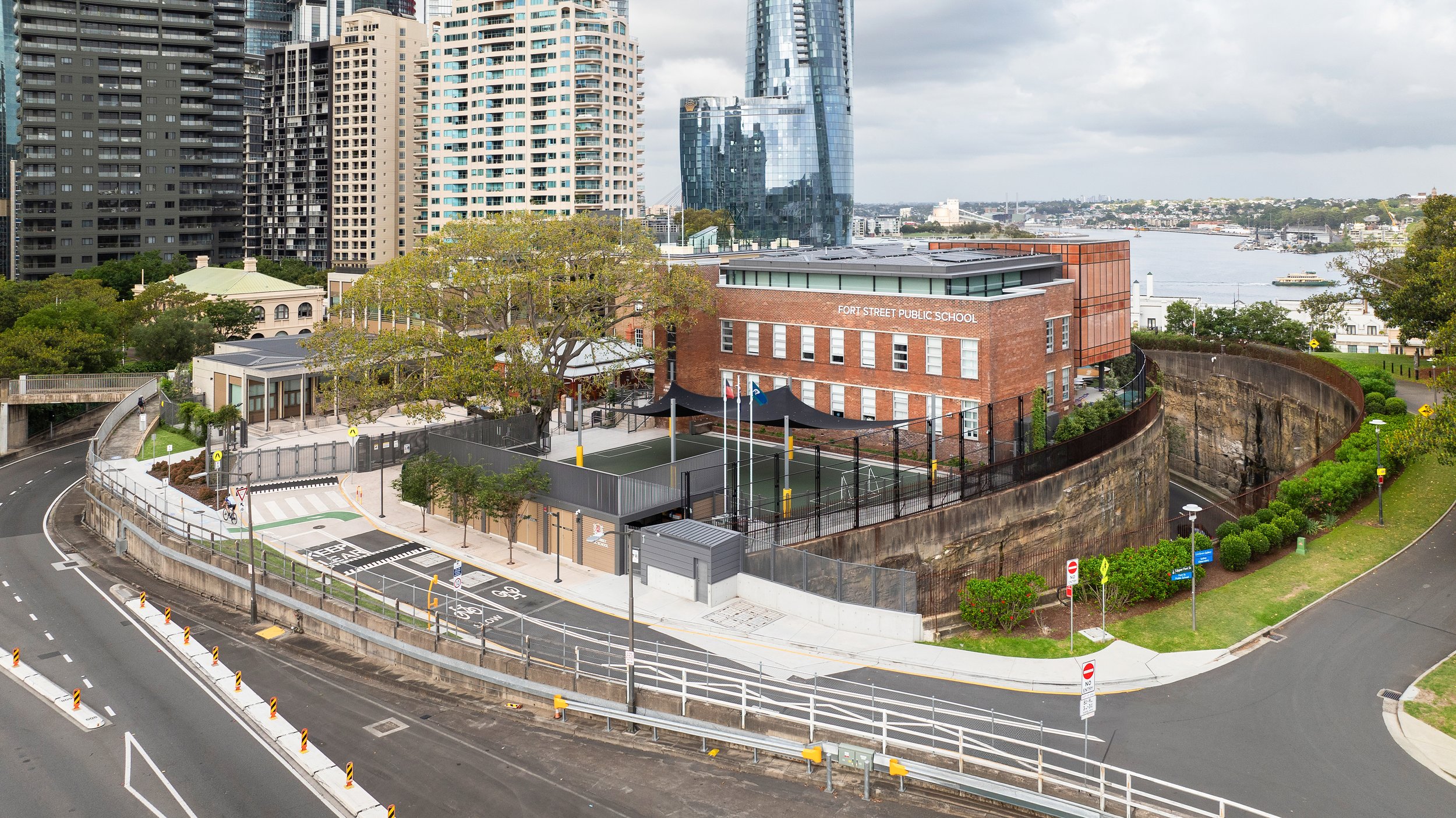 A view of Fort Street Public School in an urban area with high-rise buildings, a river in the background, and a sports court on the school grounds, surrounded by fences and trees. Designed by fjcStudio Photo by Ruth Gold