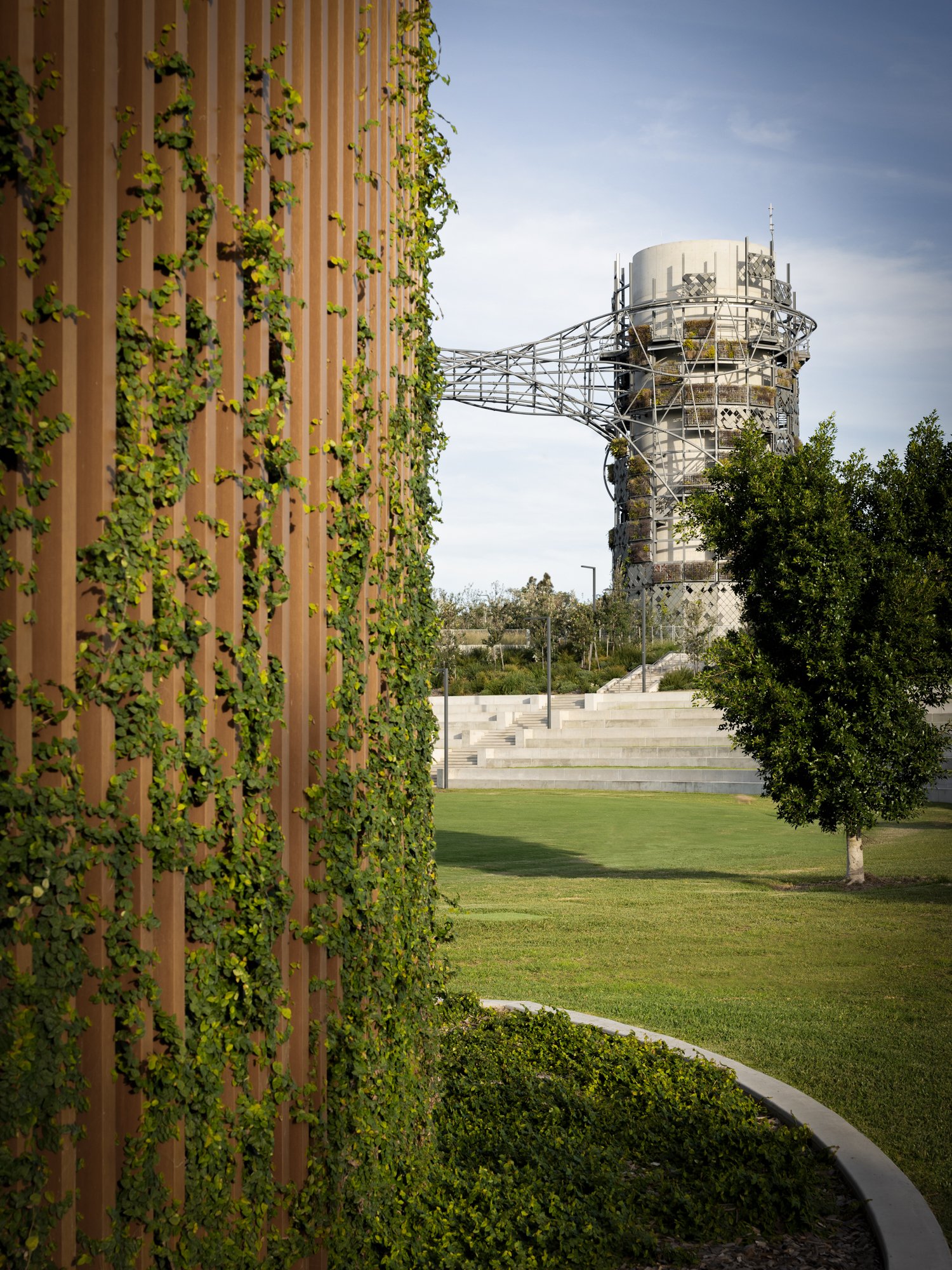 An outdoor park with a grassy lawn, a tree, and steps in the background, with a modern pipe tower connected by a bridge-like structure. Designed by Colin Polwarth Studio Photo by Ruth Gold