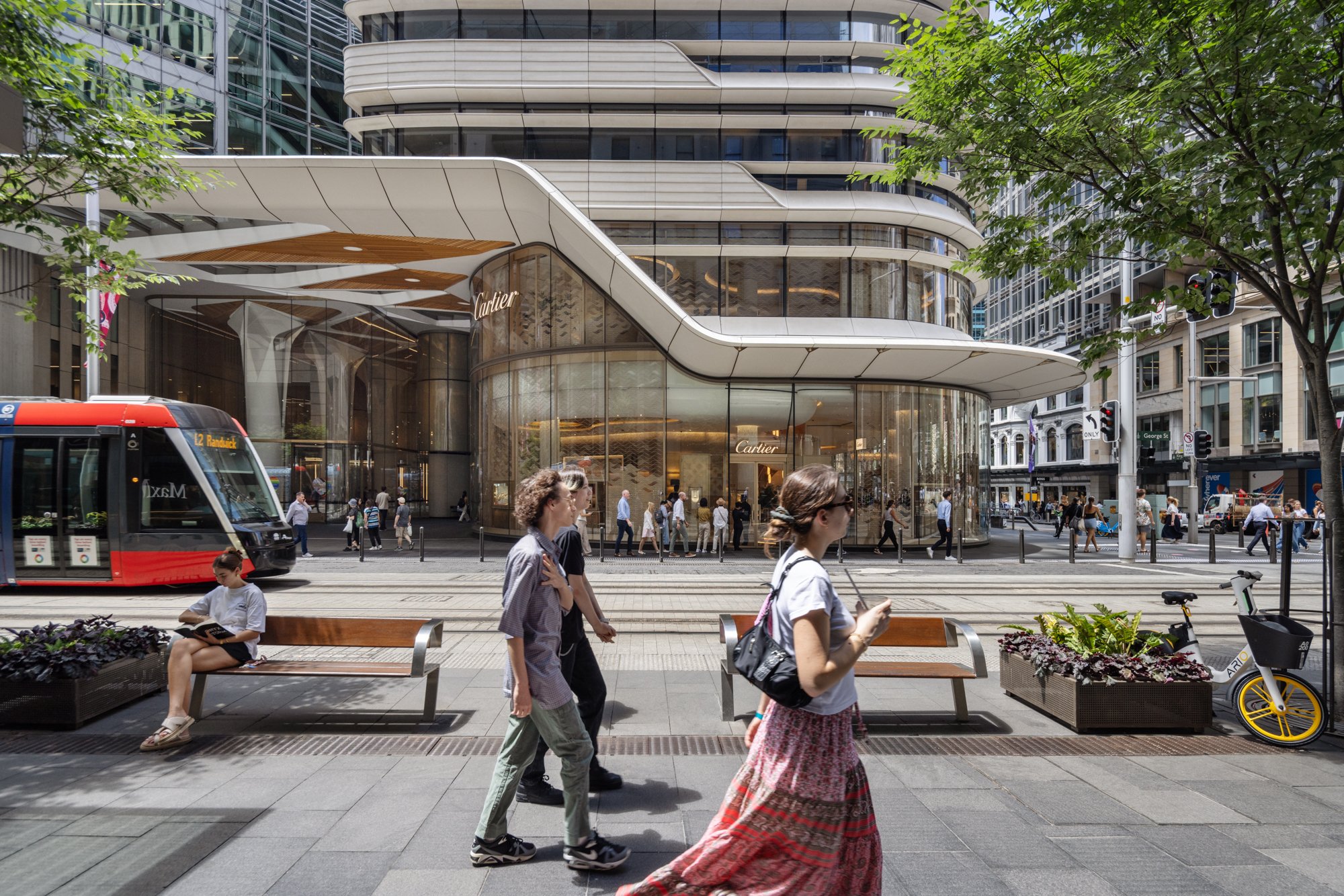 City street scene with people walking, a red tram, and a modern building with glass storefronts including a Cartier store. Designed by fjcStudio Photo by Ruth Gold