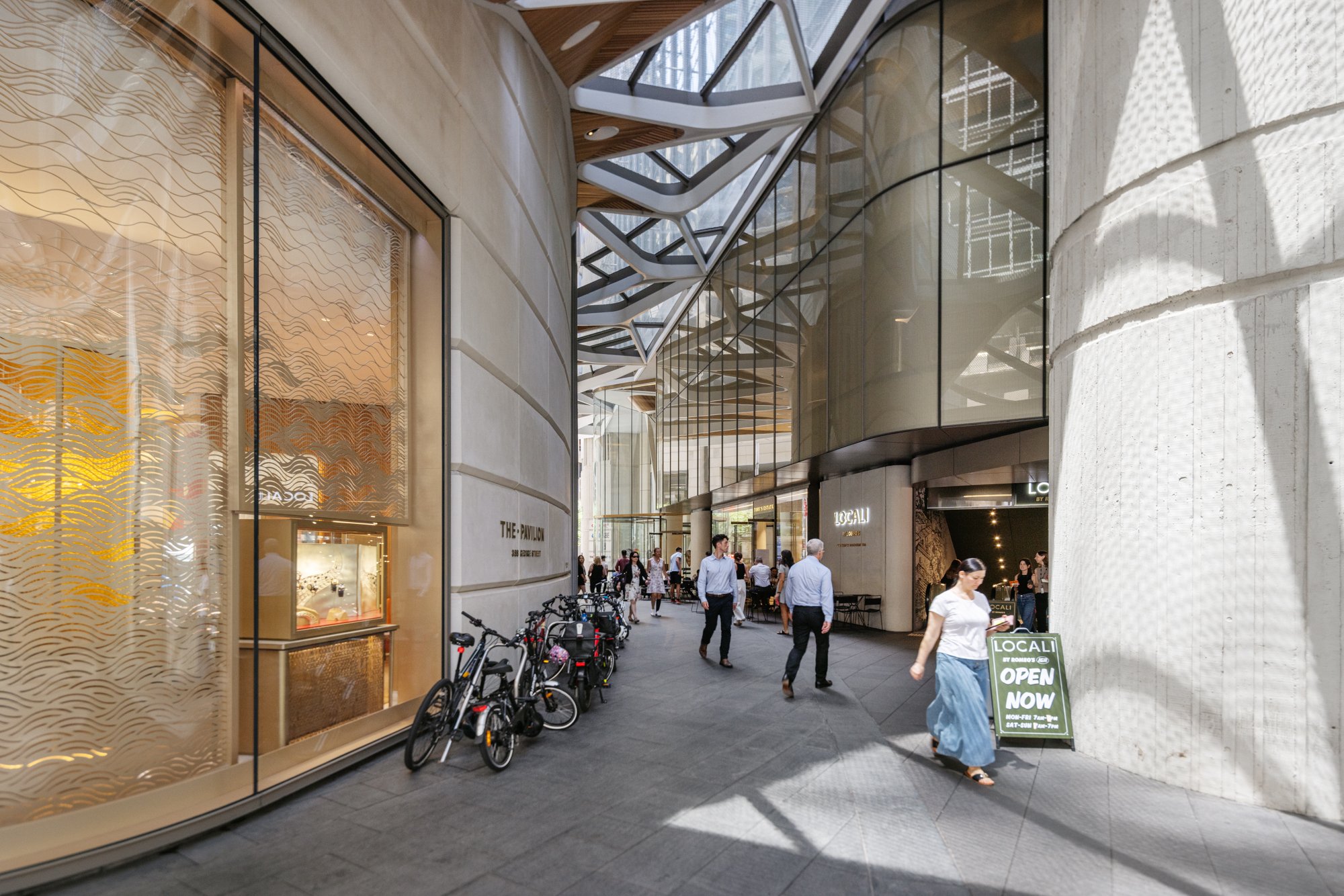 Pedestrians walking outside a modern shopping mall with a glass and metal architectural design. There are bicycles parked on the sidewalk and a signboard that says 'Open Now' in front of a store named 'Locali.' Designed by fjcStudio Photo by Ruth Gol