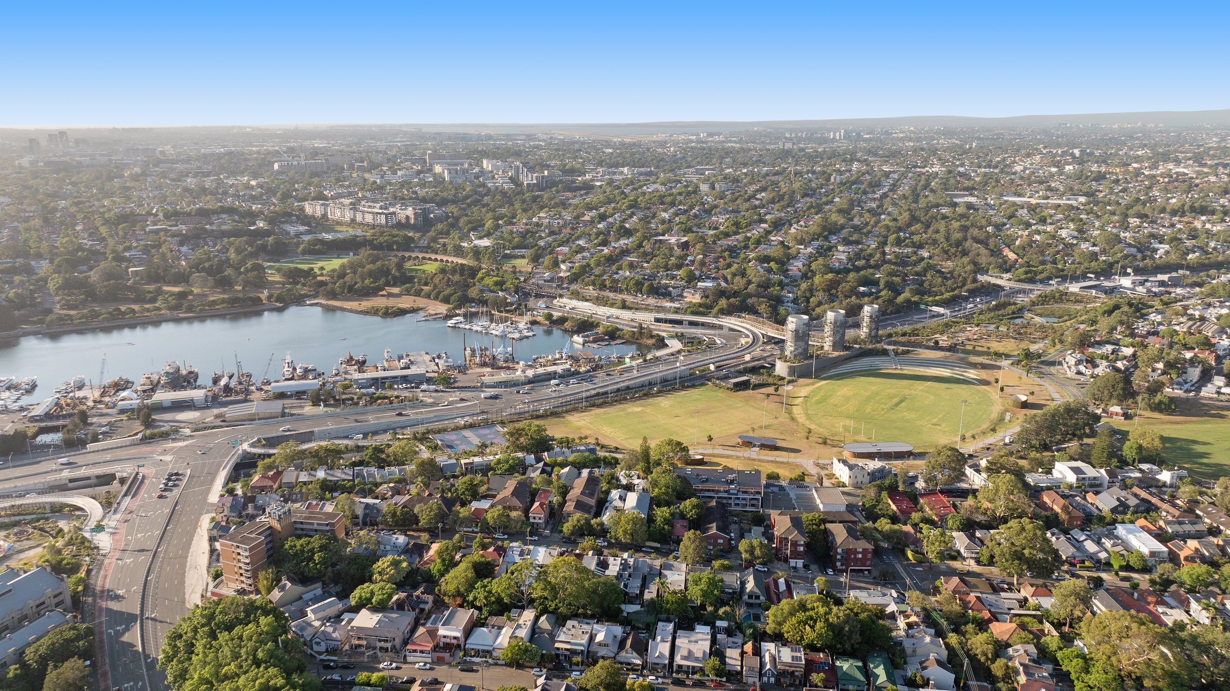 Aerial view of a city with a harbor, marina, sports field, residential neighborhoods, and green spaces under a clear blue sky. Designed by McGregor Coxall Photo by Ruth Gold