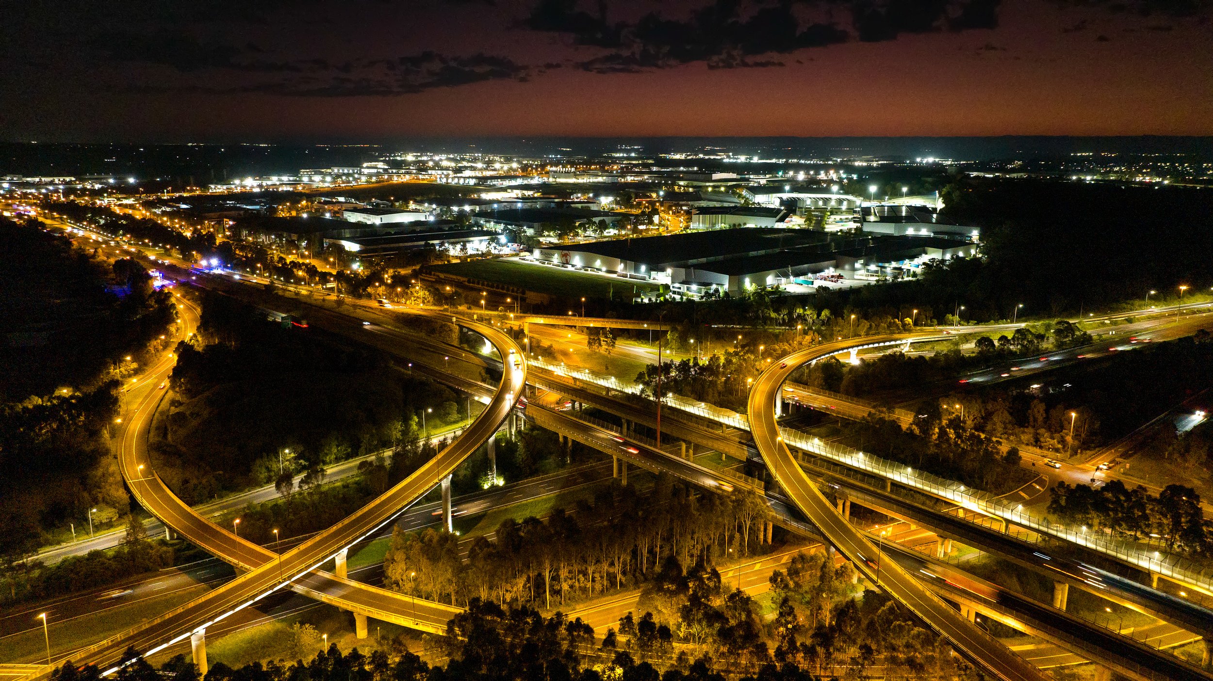 Night aerial view of an illuminated highway with multiple overpasses and curving roads in an urban area. Designed by Colin Polwarth Studio Photo by Ruth Gold