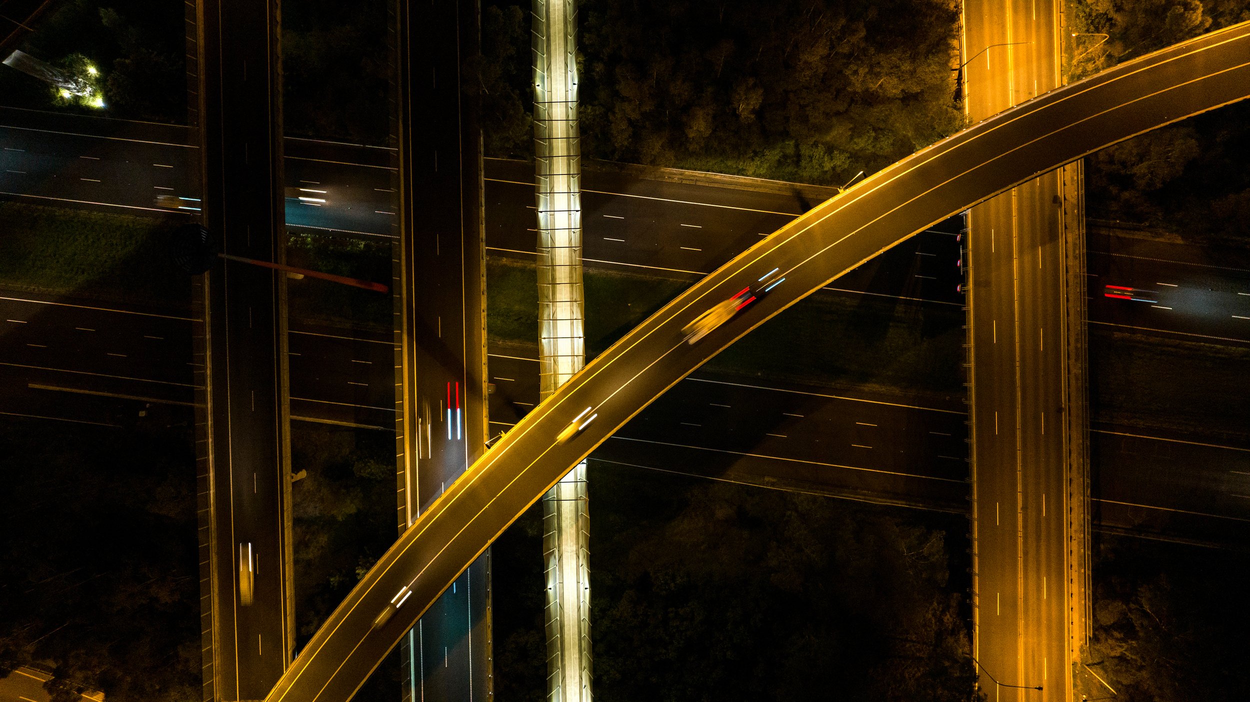 Night-time aerial view of multiple highway overpasses and roads with light trails from moving vehicles. Designed by Colin Polwarth Studio Photo by Ruth Gold